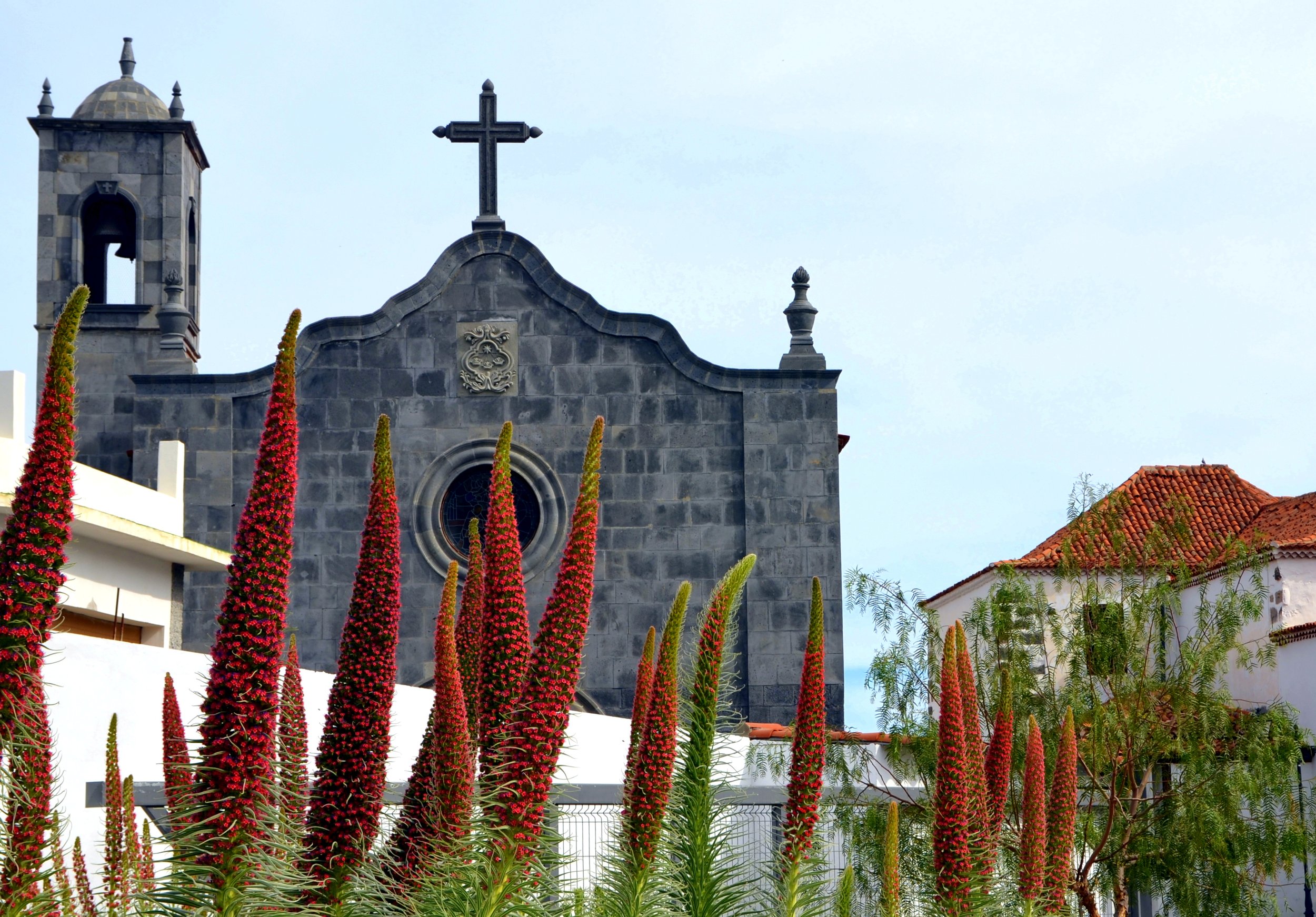 A stone church with a cross on top, surrounded by red flowering plants and white buildings with red-tiled roofs.