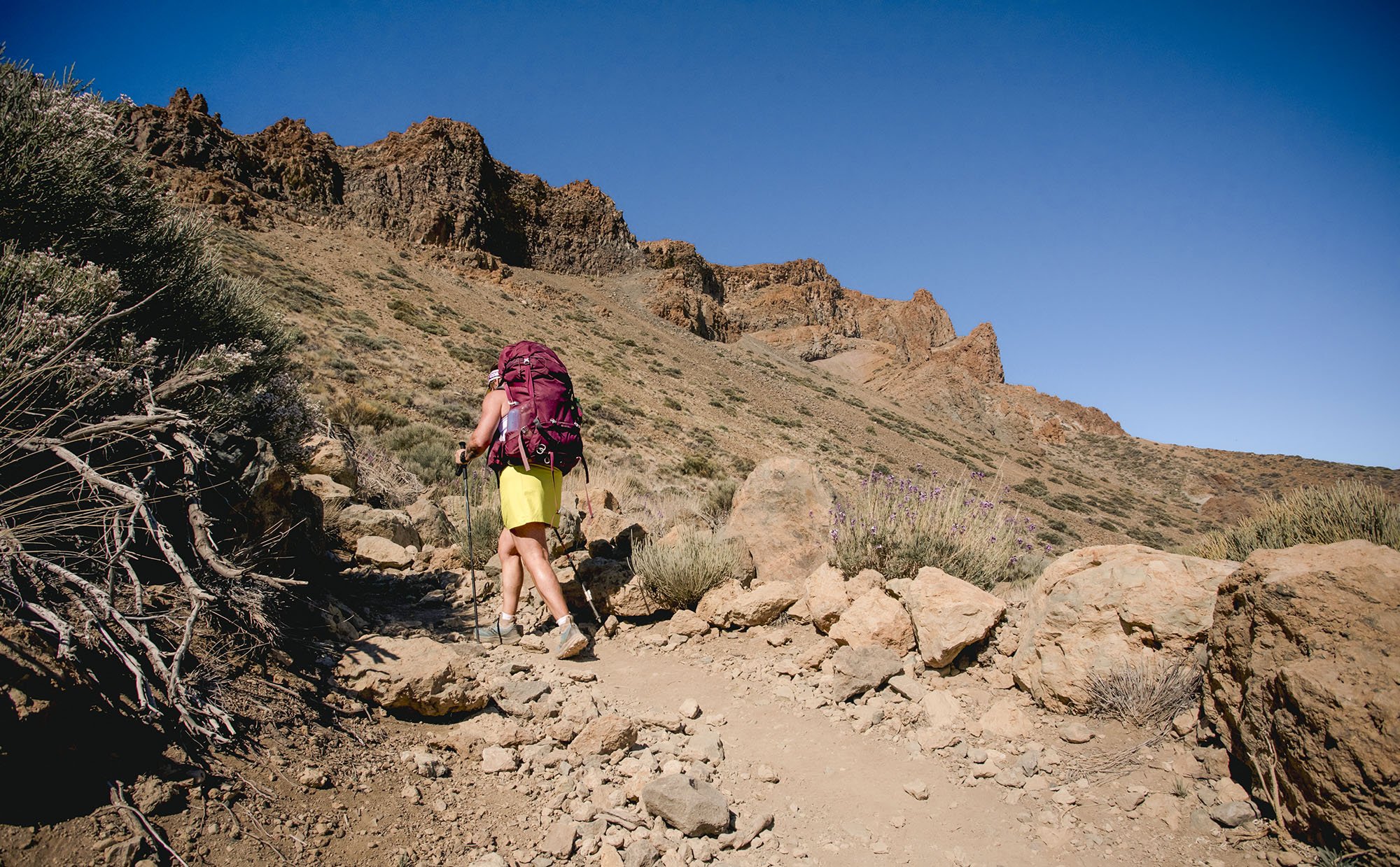 A person hiking on a rocky trail in a desert mountain landscape under a clear blue sky.