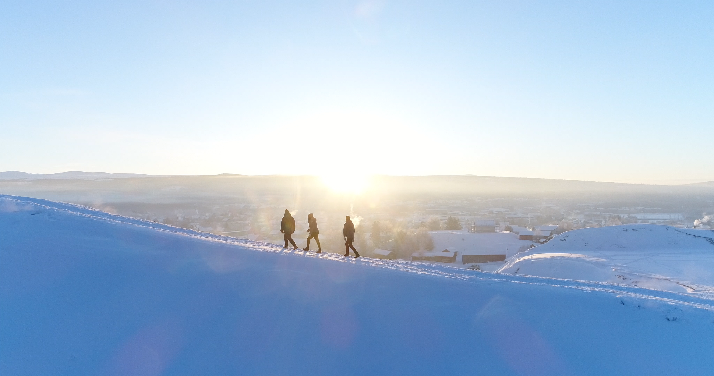 Tre personer går på snødekt landskap under solnedgang eller soloppgang, med by og fjell i bakgrunnen.