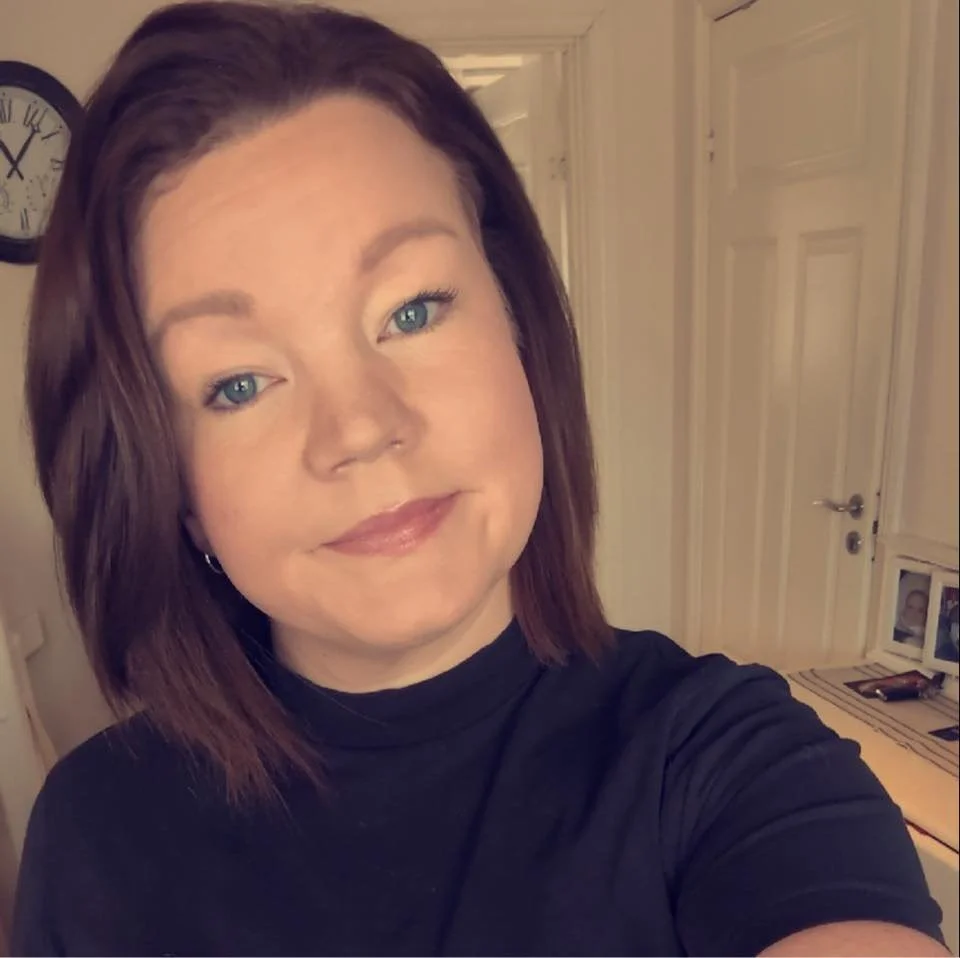 Et portrett av en kvinne med brunt hår og blå øyne som smiler, tatt i et rom med hvite vegger og en veggklokke.