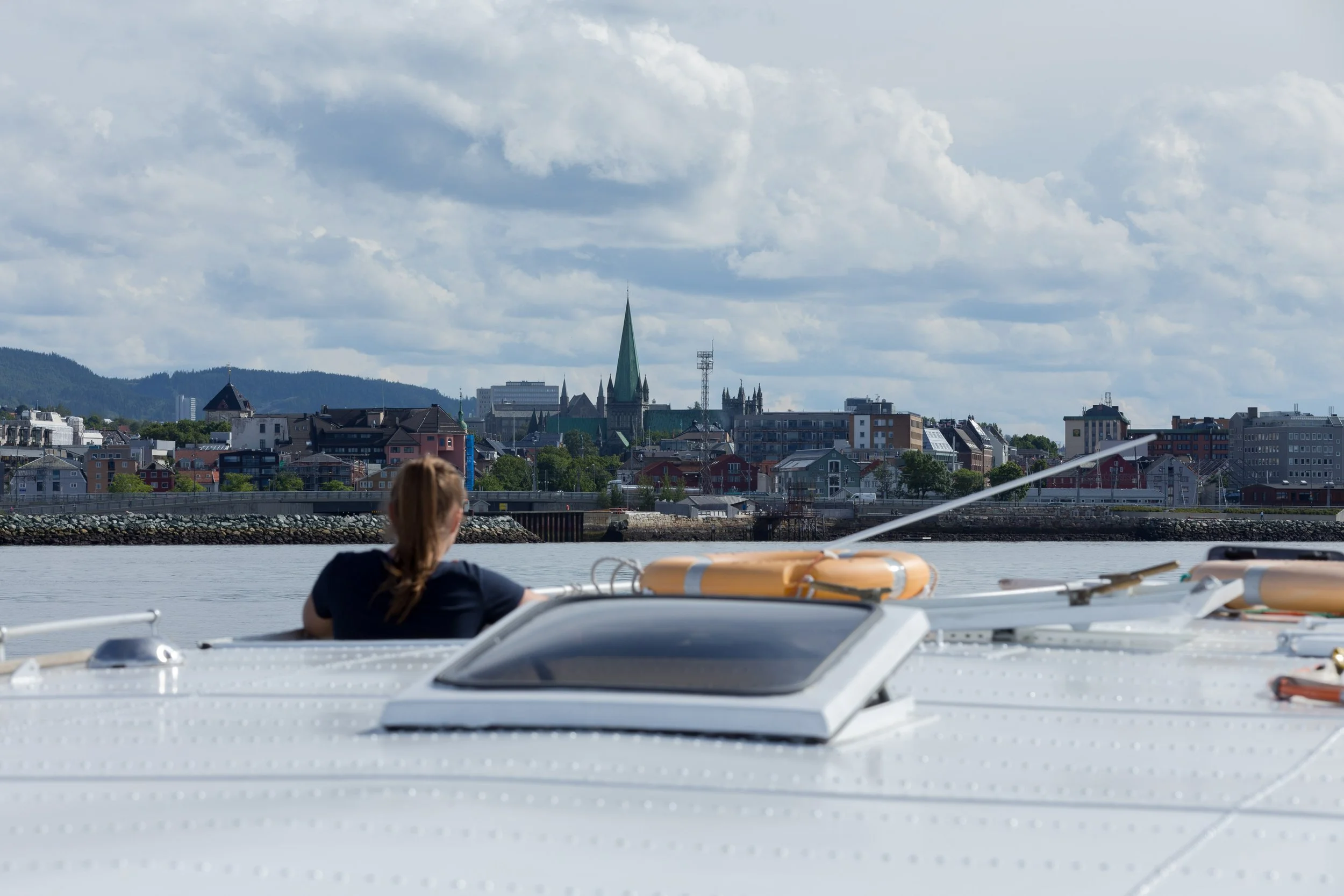 Utsikt over Trondheim by med Nidarosdomen og moderne bygninger med utsikt mot fjell i bakgrunnen, sett fra vannet hvor en person sitter i en båt.