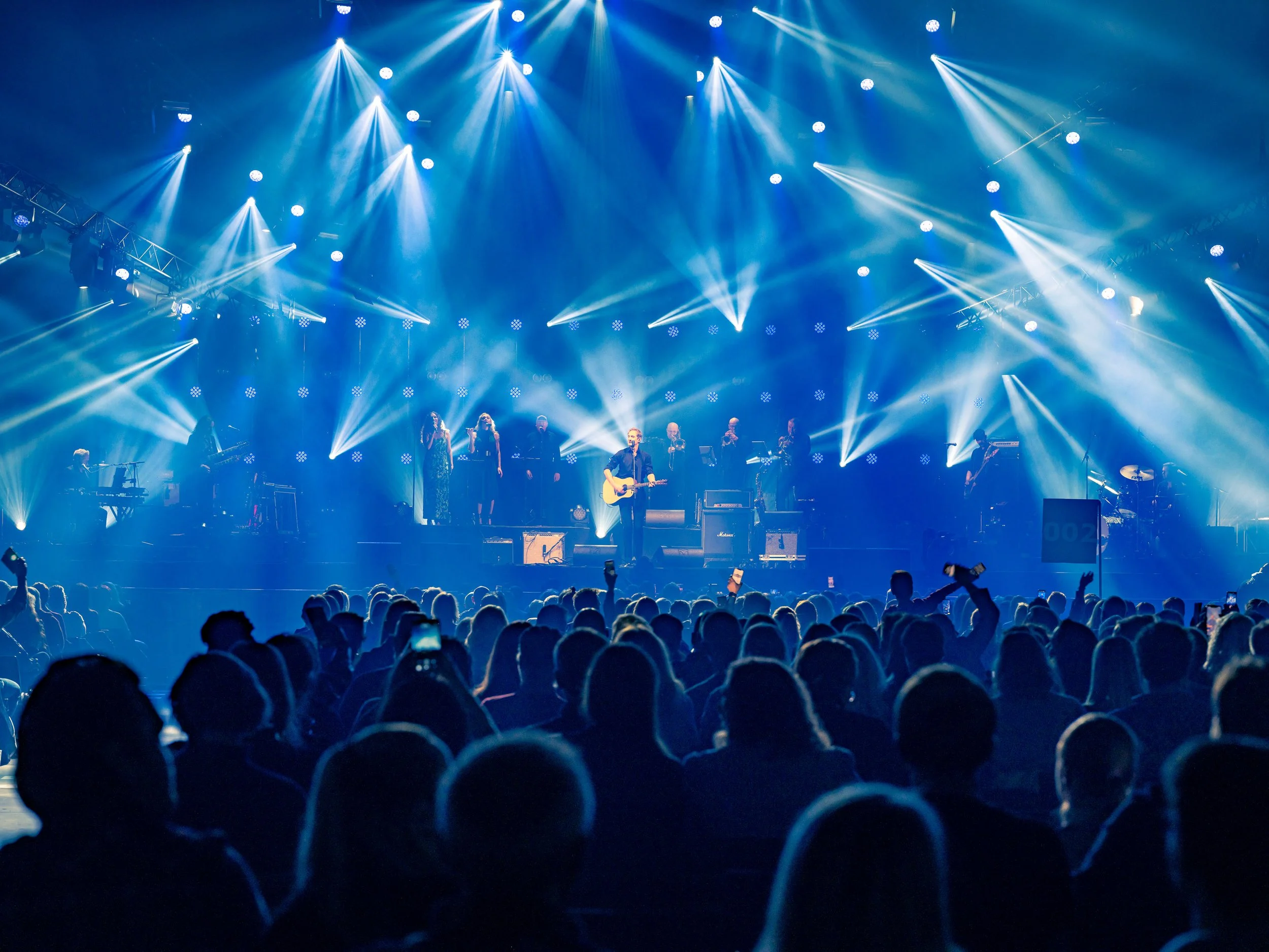 En scene med musikere under en konsert, lysende blå og hvite lys og en stor publikum i forgrunnen.