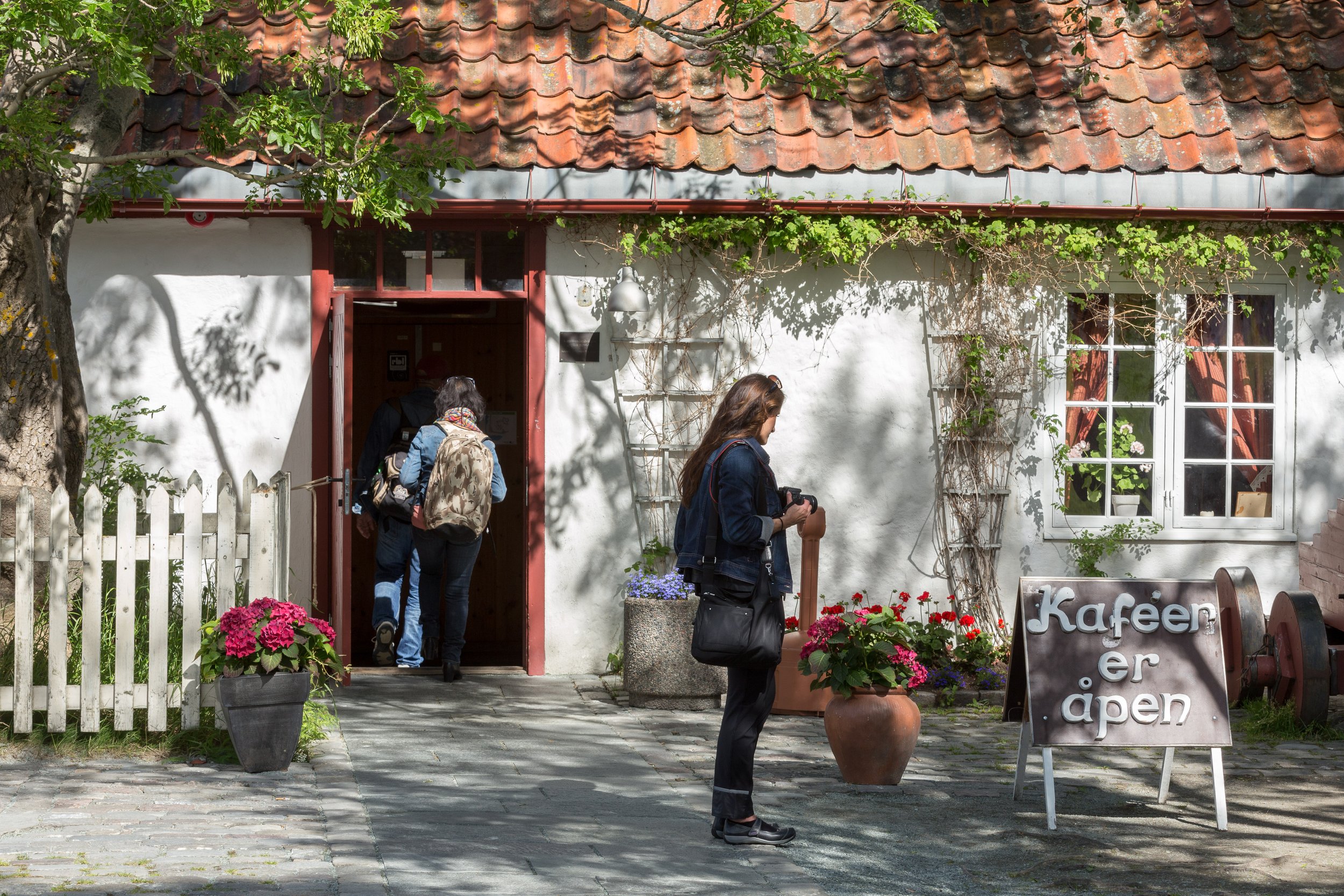 Utendørs utsikt av en liten kafé med folk som går inn og ut. Det er blomsterpotter, et gjerde, et tre og en hvit vegg med store vinduer. Takhengene er av mørke tegler, og det er skygger av tre i forgrunnen.Kafe og selskapslokaler på Munkholmen.