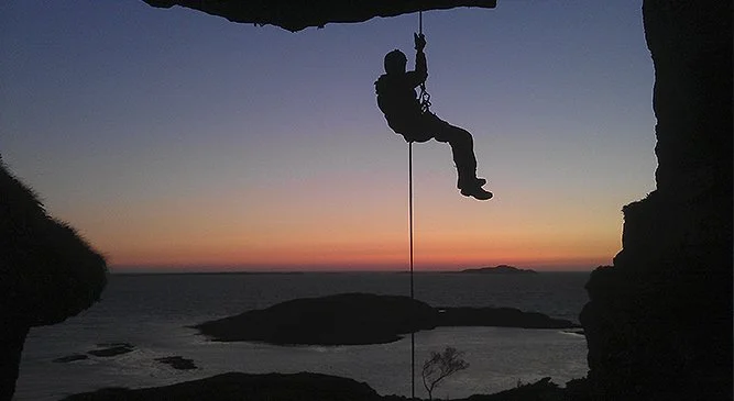 En person som klatrer opp en fjellvegg ved solnedgang, hengende i zip-line. Silhuett mot en farget himmel.