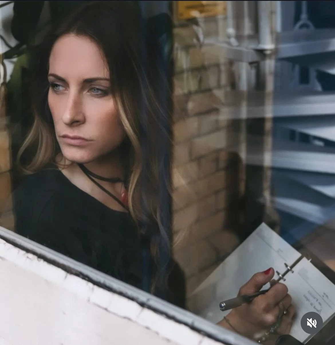 A woman with dark hair and light skin looking out a window, holding a pen and paper, with a brick wall and metal structures in the background.