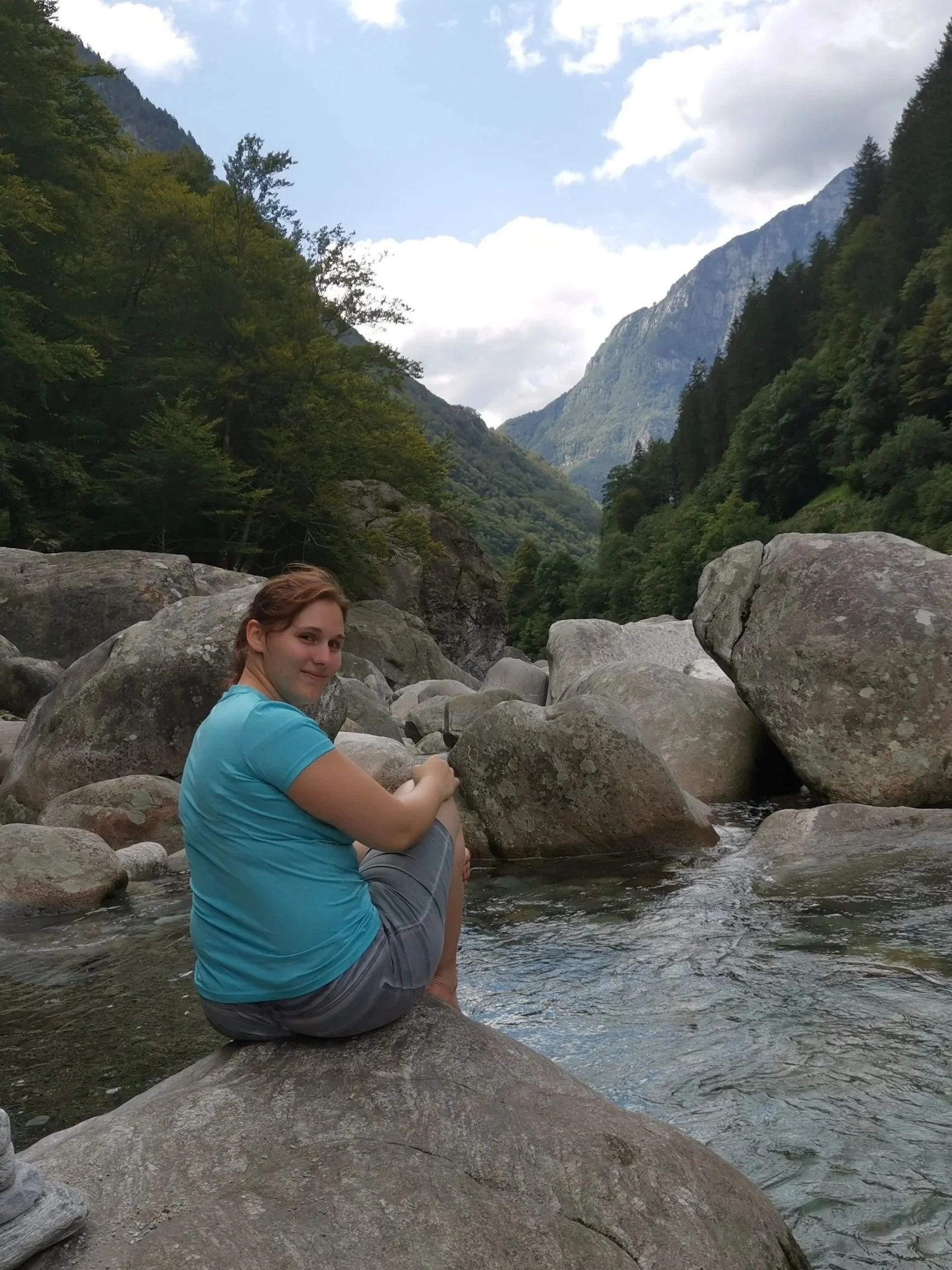 Junge Frau sitzt auf einem großen Stein im Fluss in einer Berglandschaft mit Bäumen und Wolken am Himmel.