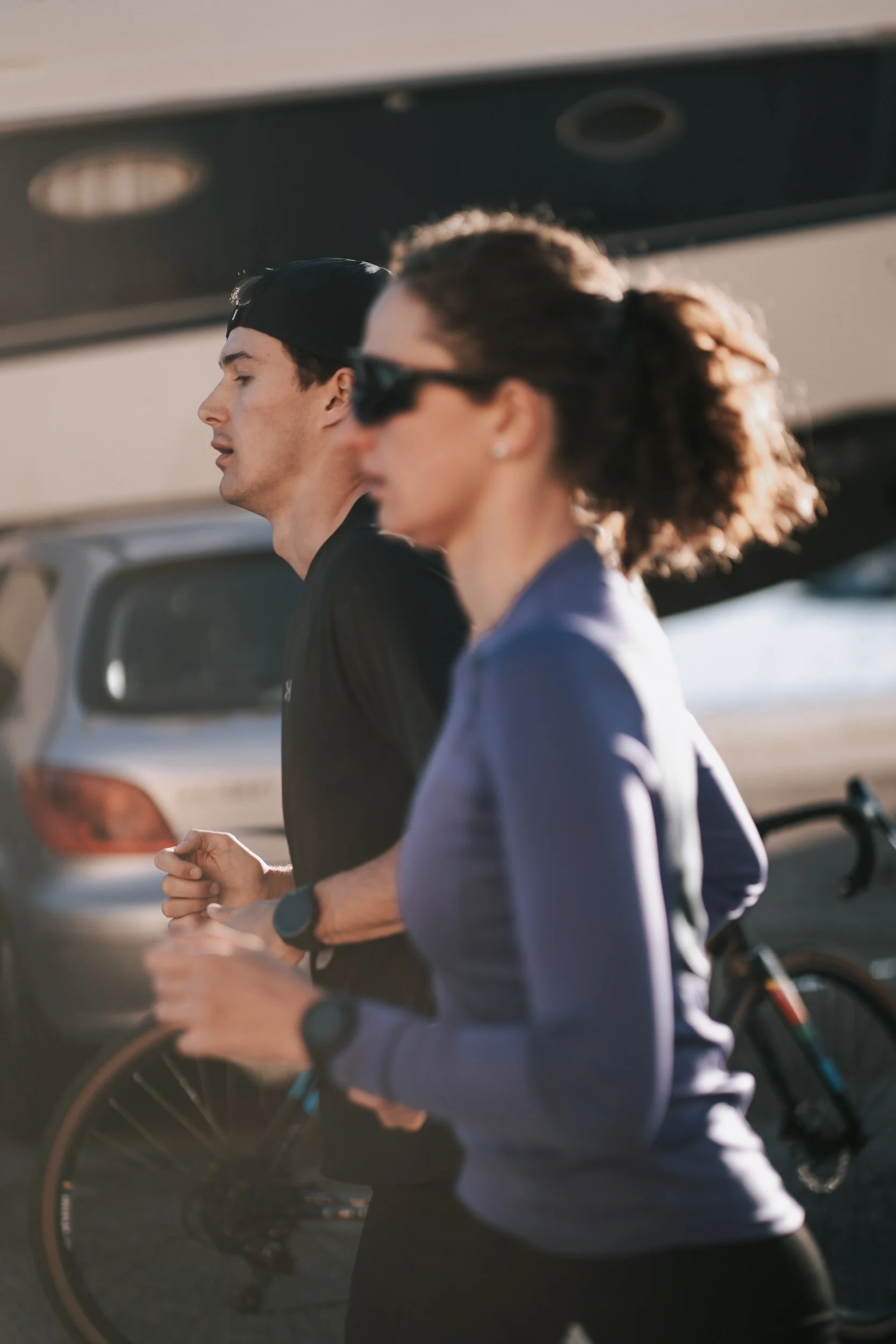 Two people jogging outdoors near parked cars, one woman wearing sunglasses and a blue jacket, and one man wearing a black cap and a black shirt, both with fitness trackers on their wrists.