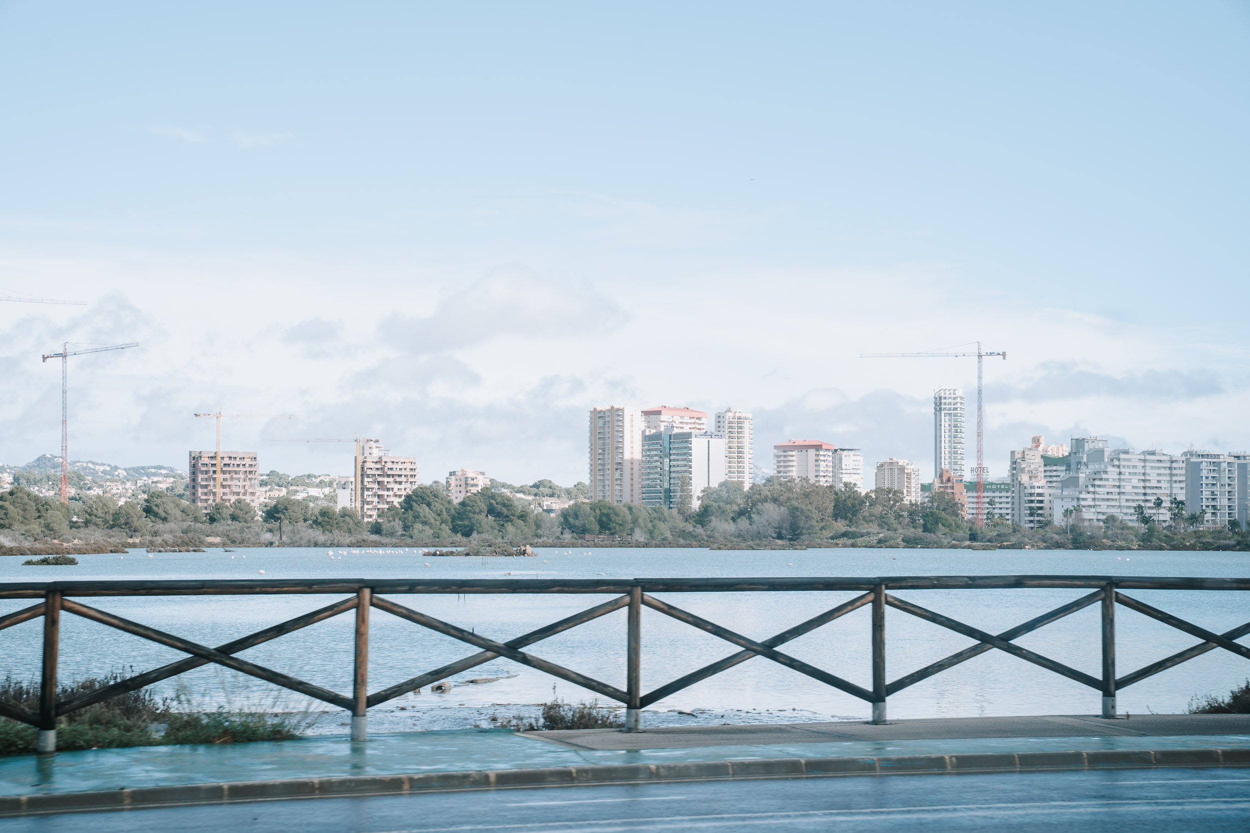 City skyline with tall buildings, some under construction with cranes, across a calm body of water, with a wooden fence in the foreground and a partly cloudy sky overhead.