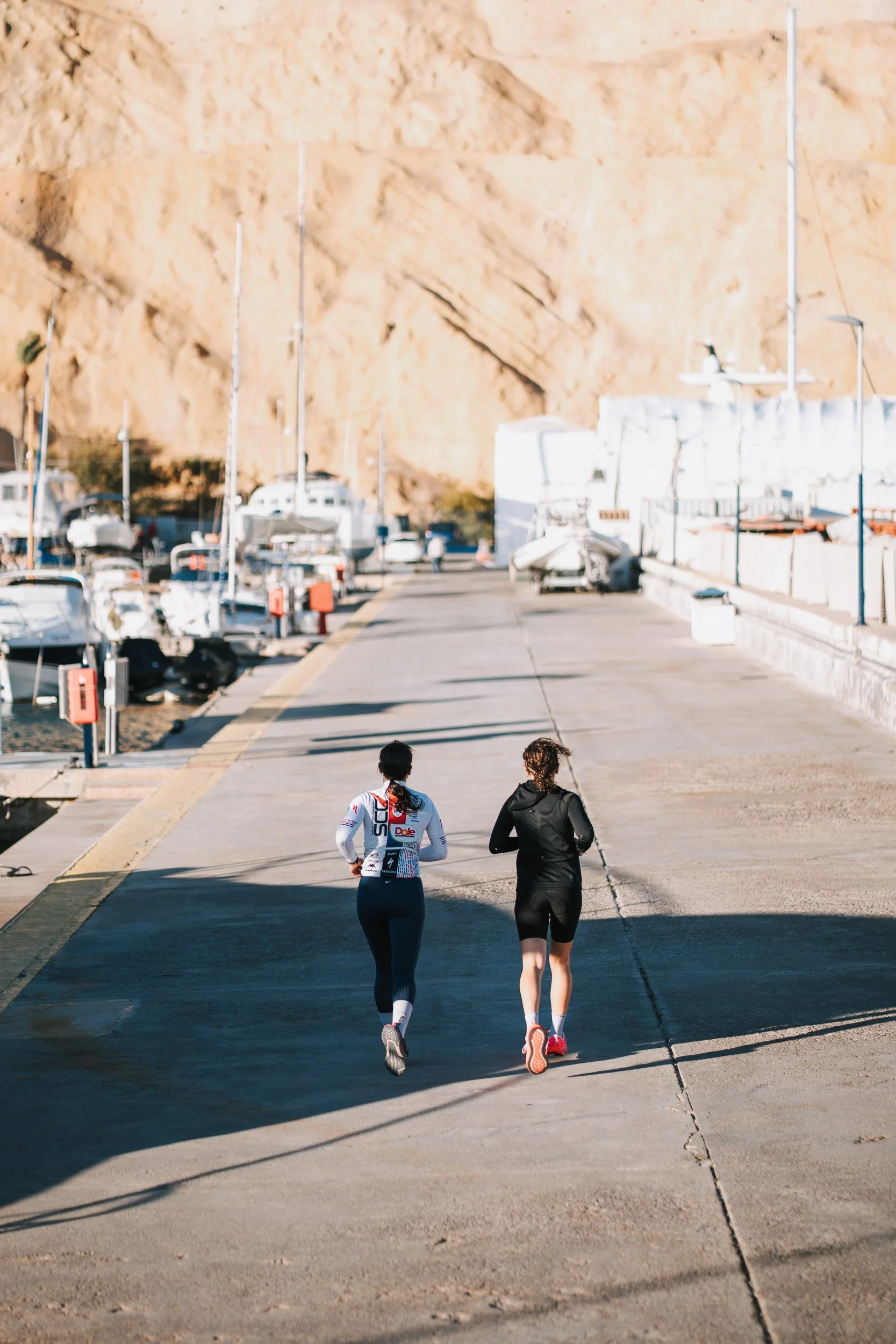 Two women jogging along a harbor with boats, dock, and orange rock formations in the background.