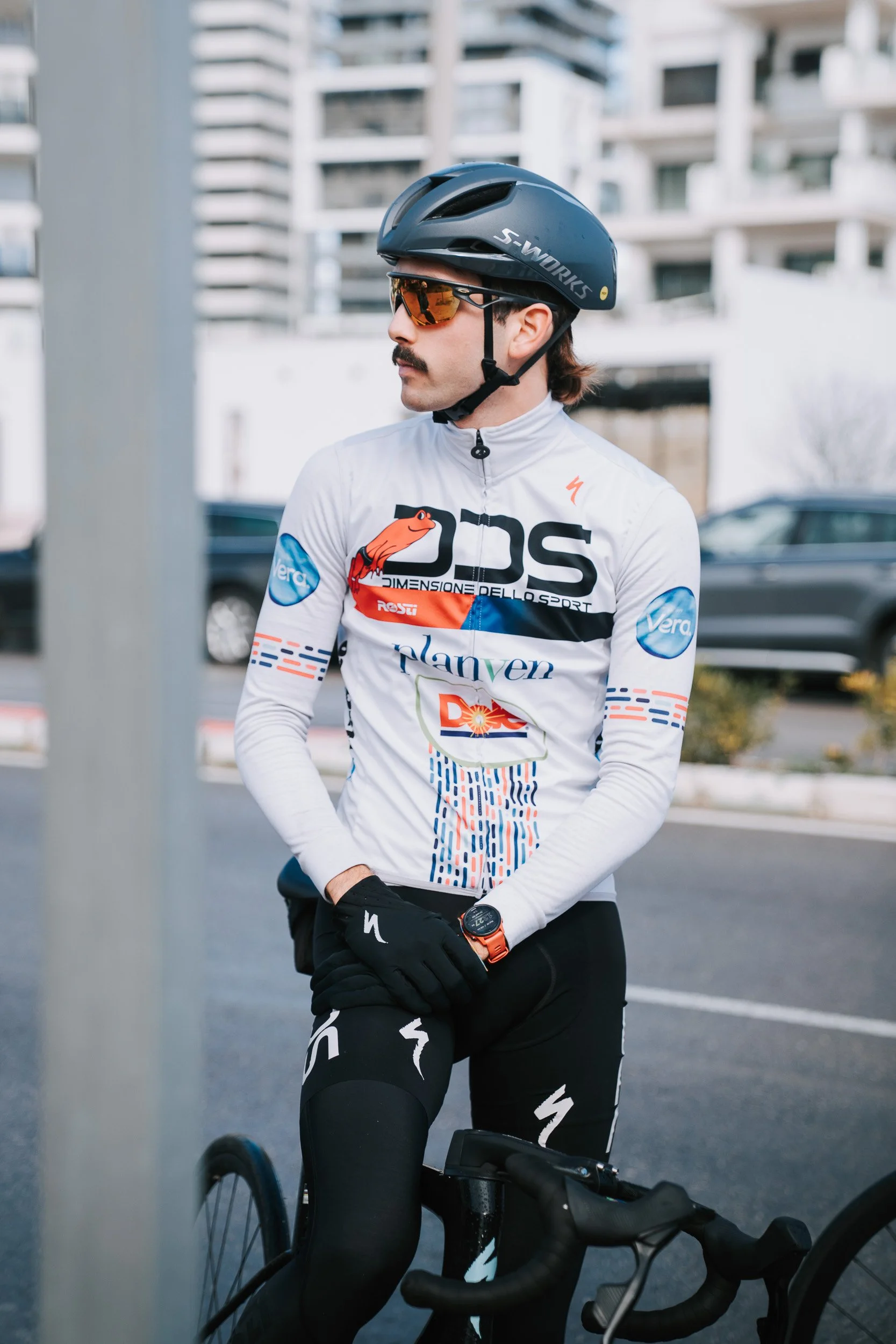 A man dressed in cycling gear, wearing a helmet, sunglasses, and gloves, standing next to a bicycle on a city street.