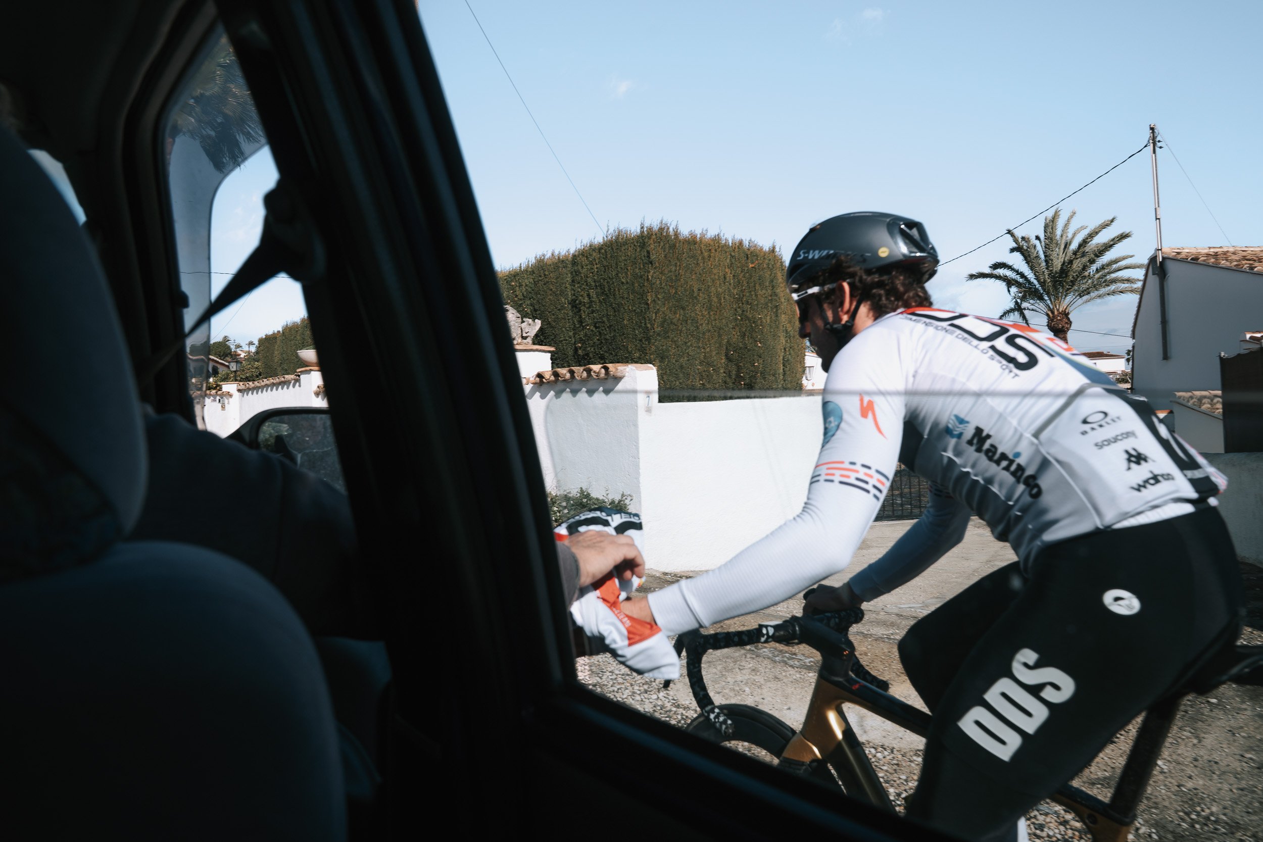 Cyclist wearing a white and black jersey and black shorts, riding a bike, and receiving a hand from someone inside a vehicle, with a white wall, trees, and utility poles in the background.