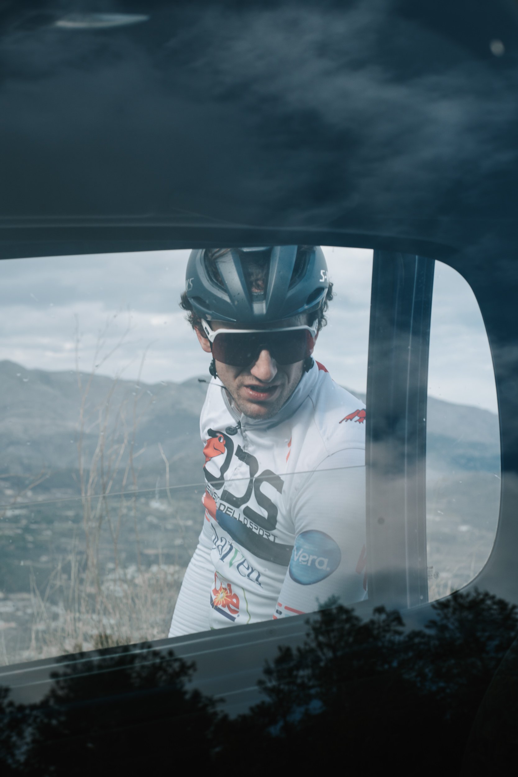 Cyclist wearing a black helmet and sunglasses looking through a car window with a landscape of mountains and cloudy sky in the background.