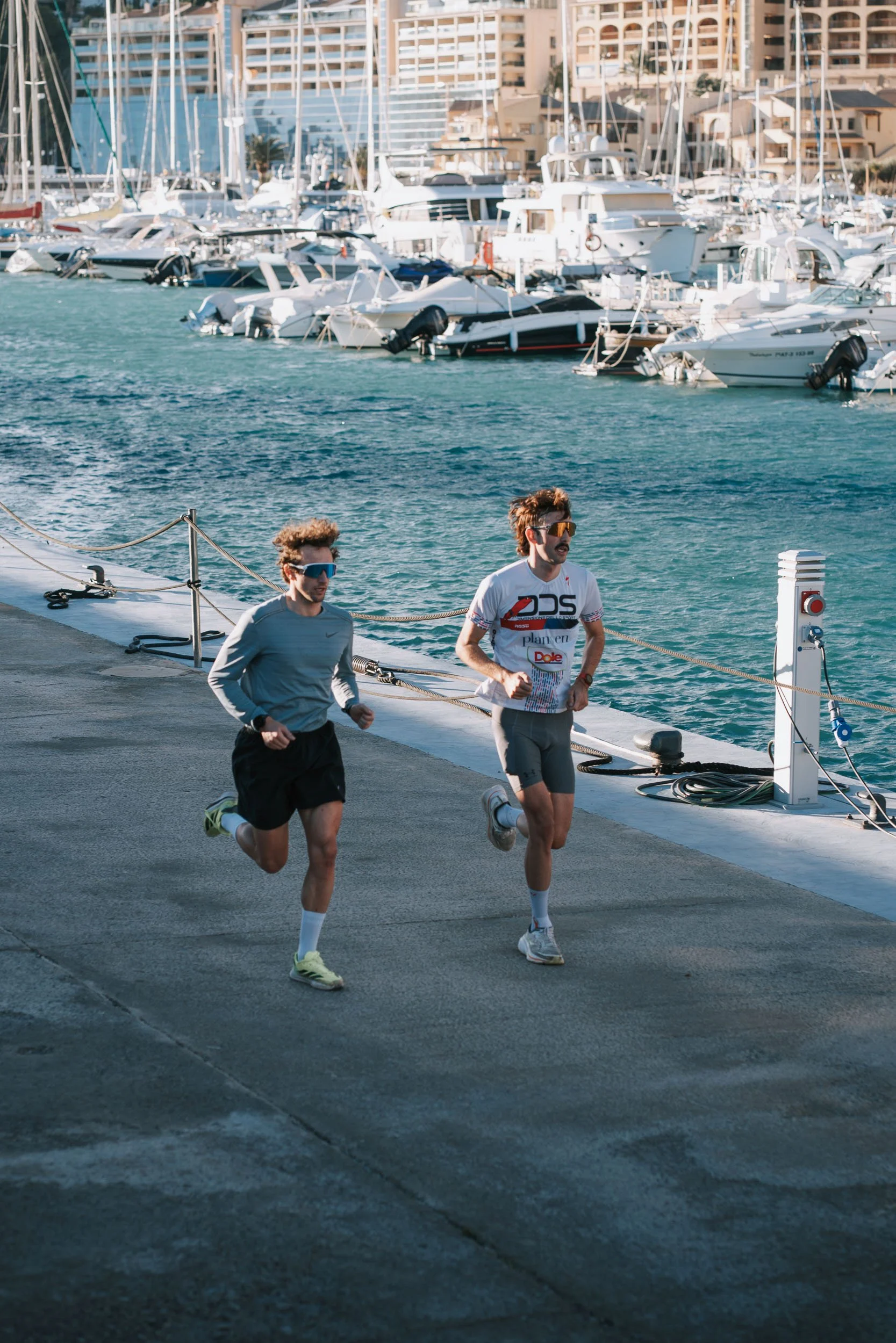 Two men running on a dock near a marina with boats and yachts in the background.