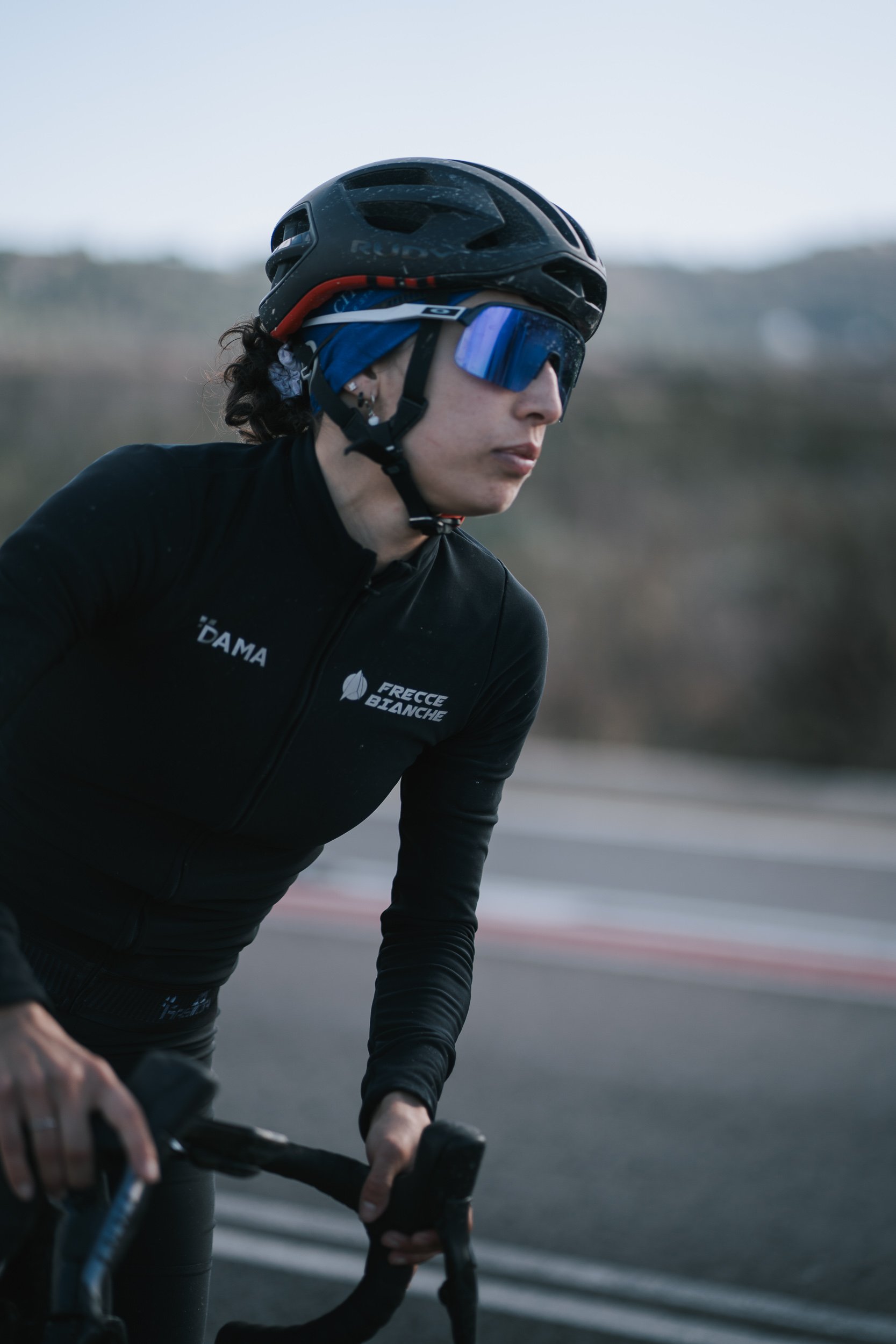 A female cyclist wearing a black helmet, sunglasses, and a black cycling outfit riding on a road.