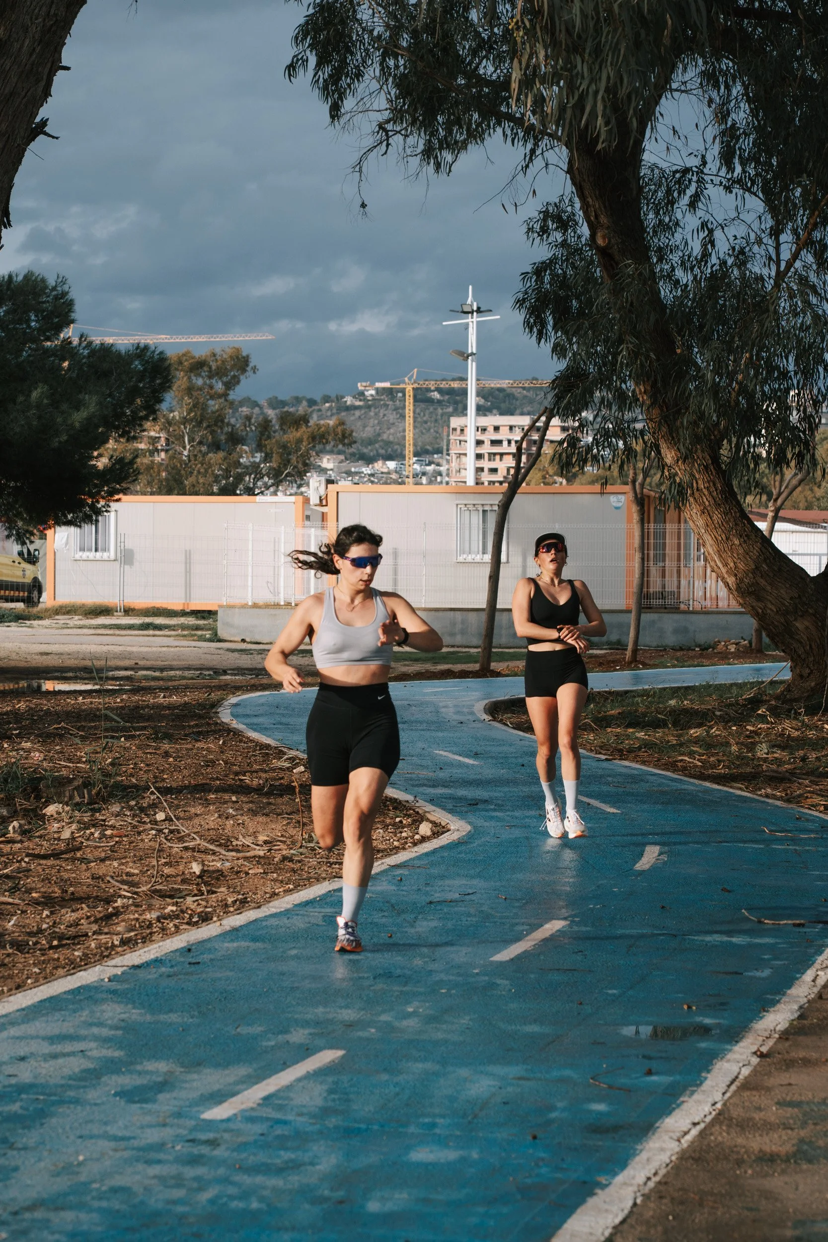Two women jogging on a blue running track outdoors, surrounded by trees, with buildings and a cloudy sky in the background.