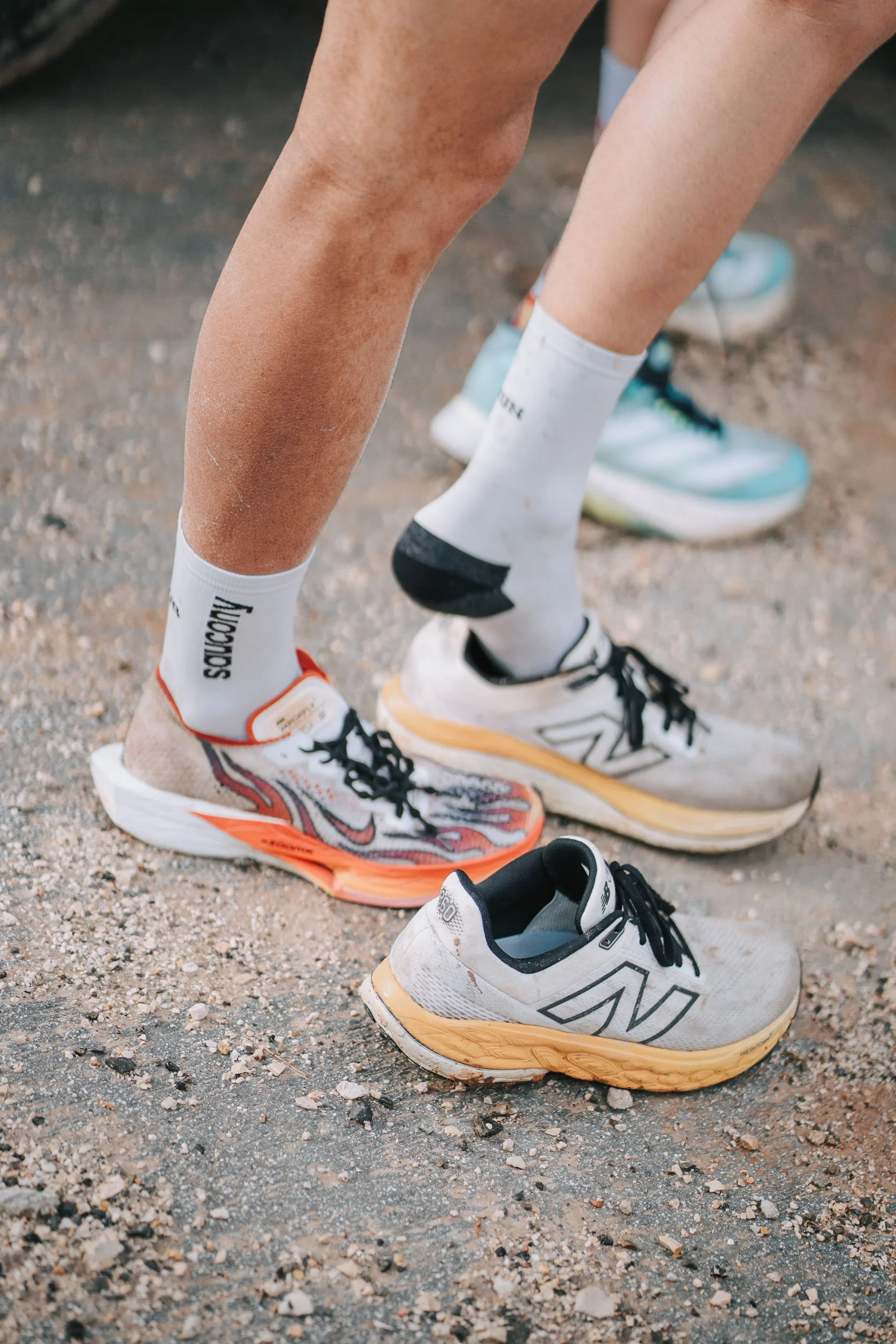 Close-up of a feet, with one foot in colorful running sneakers and the other foot with a plain sneaker, on a gravel surface. The person is wearing white socks.