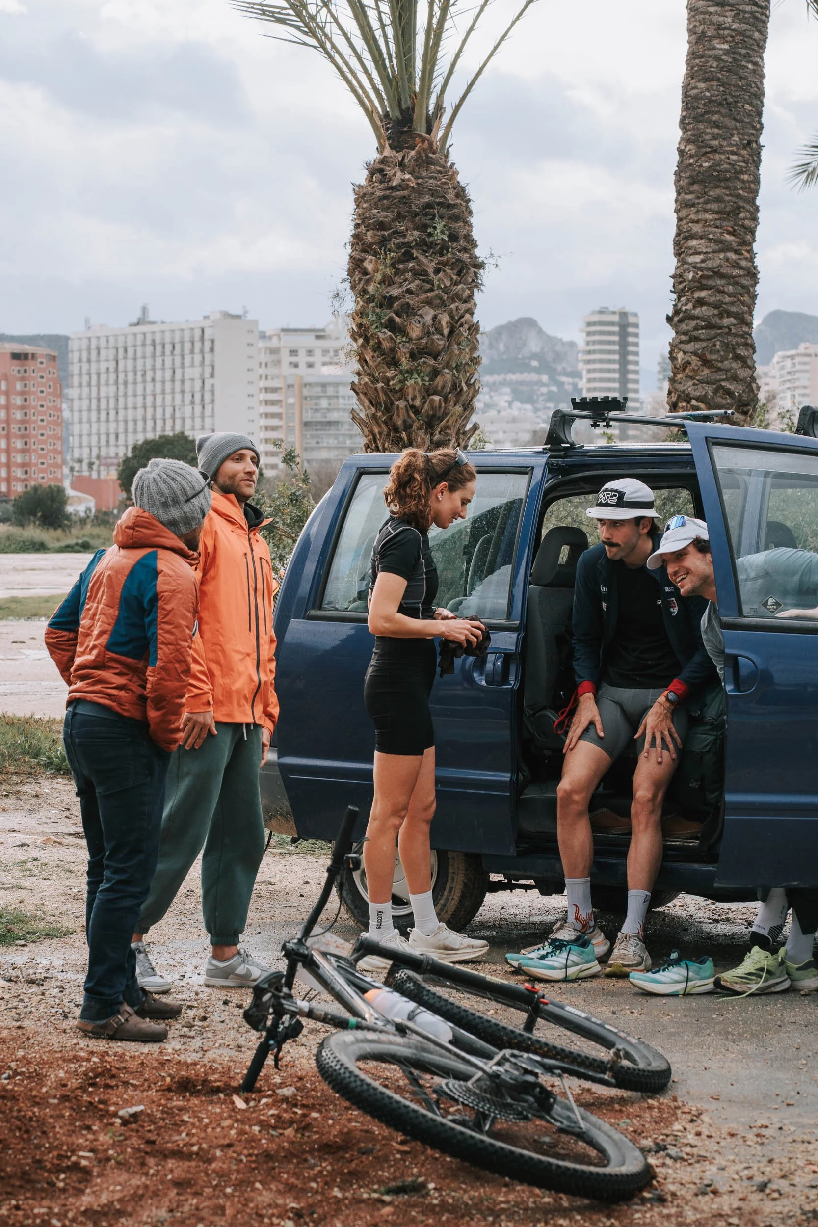 Group of five people, dressed in casual outdoor gear, preparing for biking adventure by a blue van with two palm trees in the background. One person is sitting inside the van, others are standing nearby, and a mountain bike is on the ground.