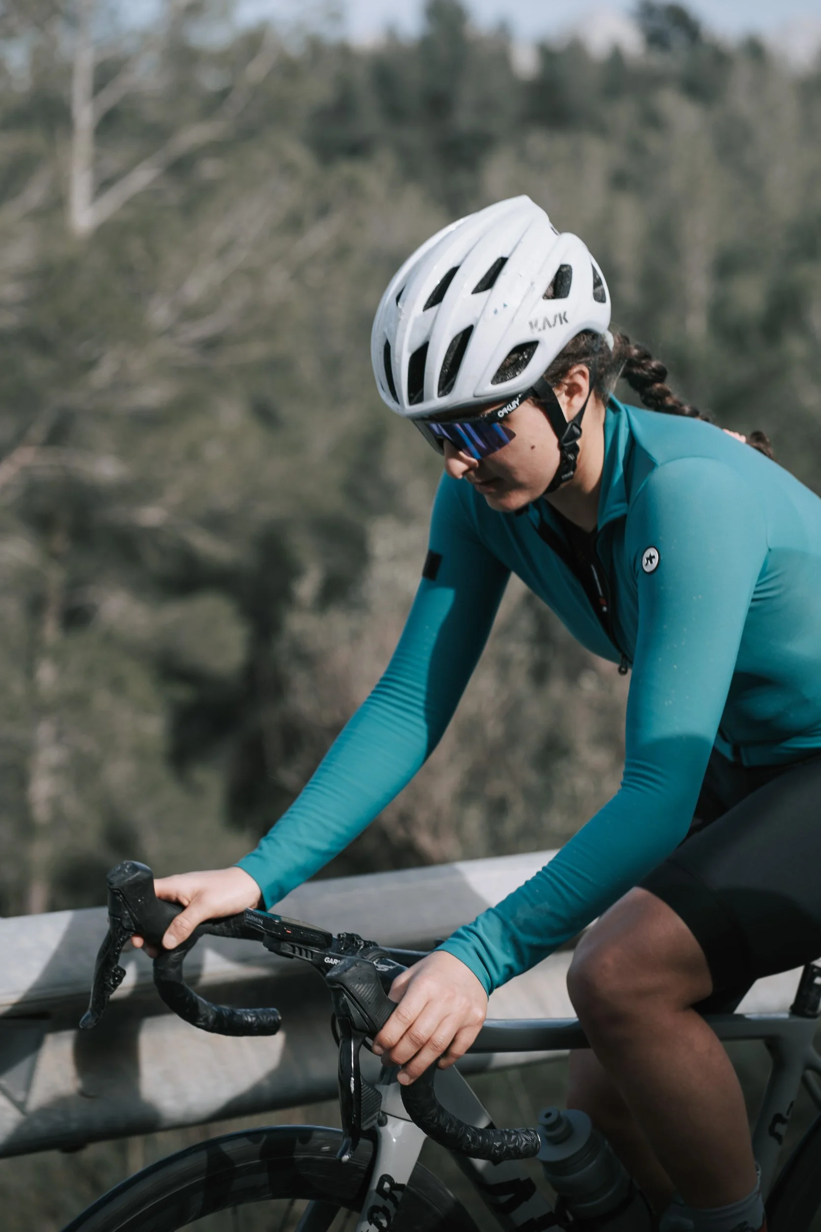 A female cyclist wearing a white helmet, sunglasses, a teal long-sleeve cycling jersey, and black shorts riding a road bike outdoors on a trail with trees in the background.