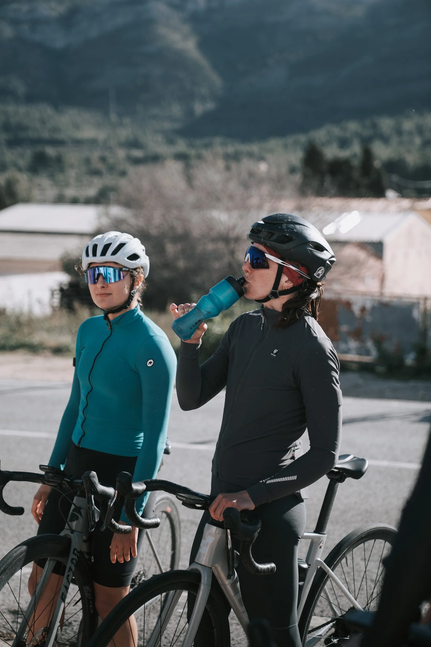 Two women in cycling gear, wearing helmets and sunglasses, taking a break with their bikes in a rural area; one woman drinking from a water bottle.
