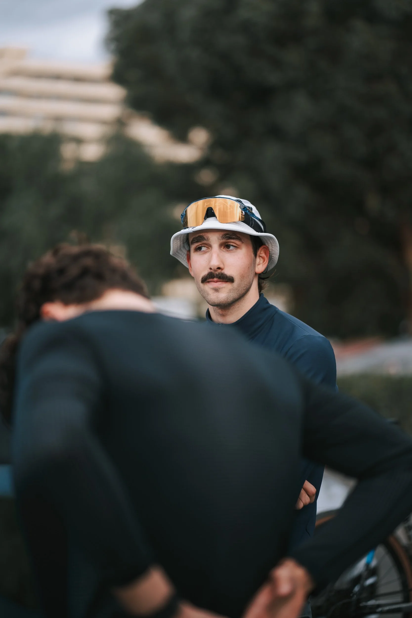A man with a mustache wearing a white bucket hat and orange-tinted sunglasses on top, looking thoughtfully at another person, outdoors with trees and a building in the background.