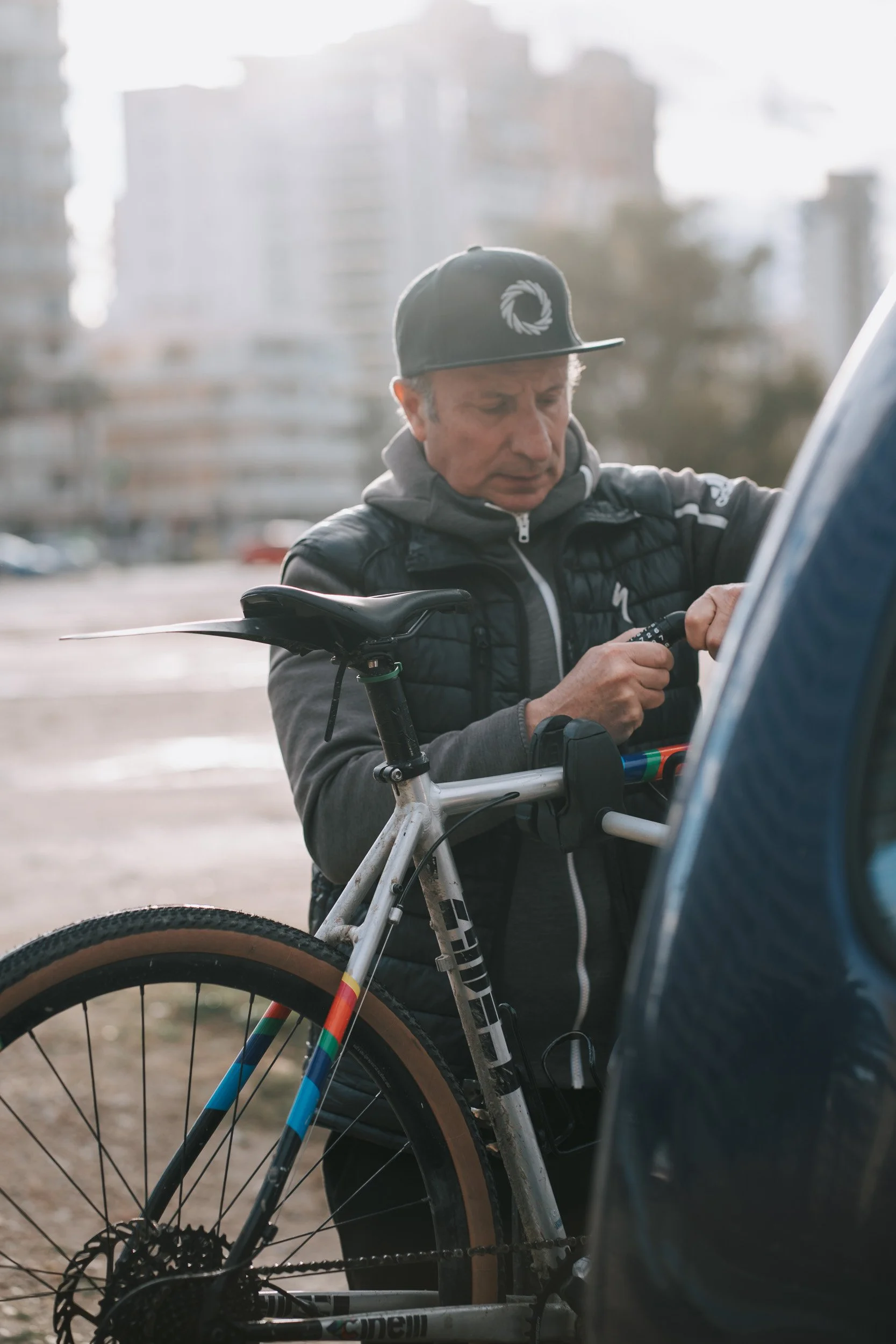 A man wearing a black cap, gray hoodie, and black vest working on his bicycle next to a car in an outdoor setting, with blurry buildings in the background.