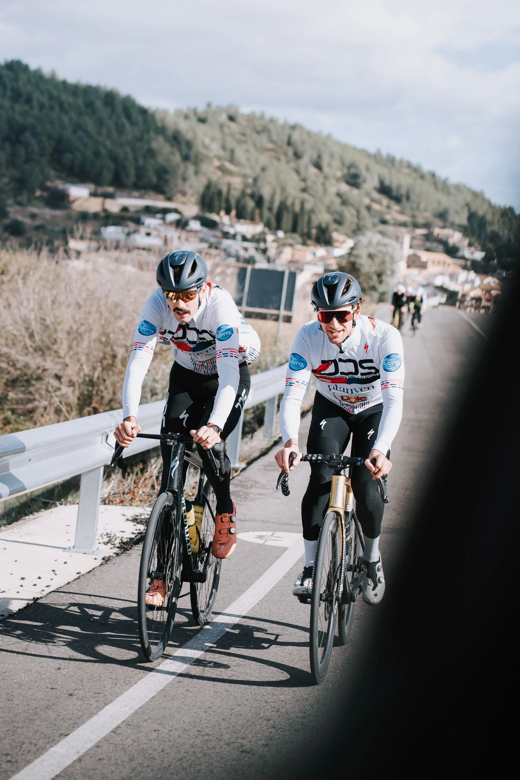 Two male cyclists riding their bikes uphill on a mountain road with a guardrail, wearing helmets and cycling jerseys, with a scenic background of hills and a small town.