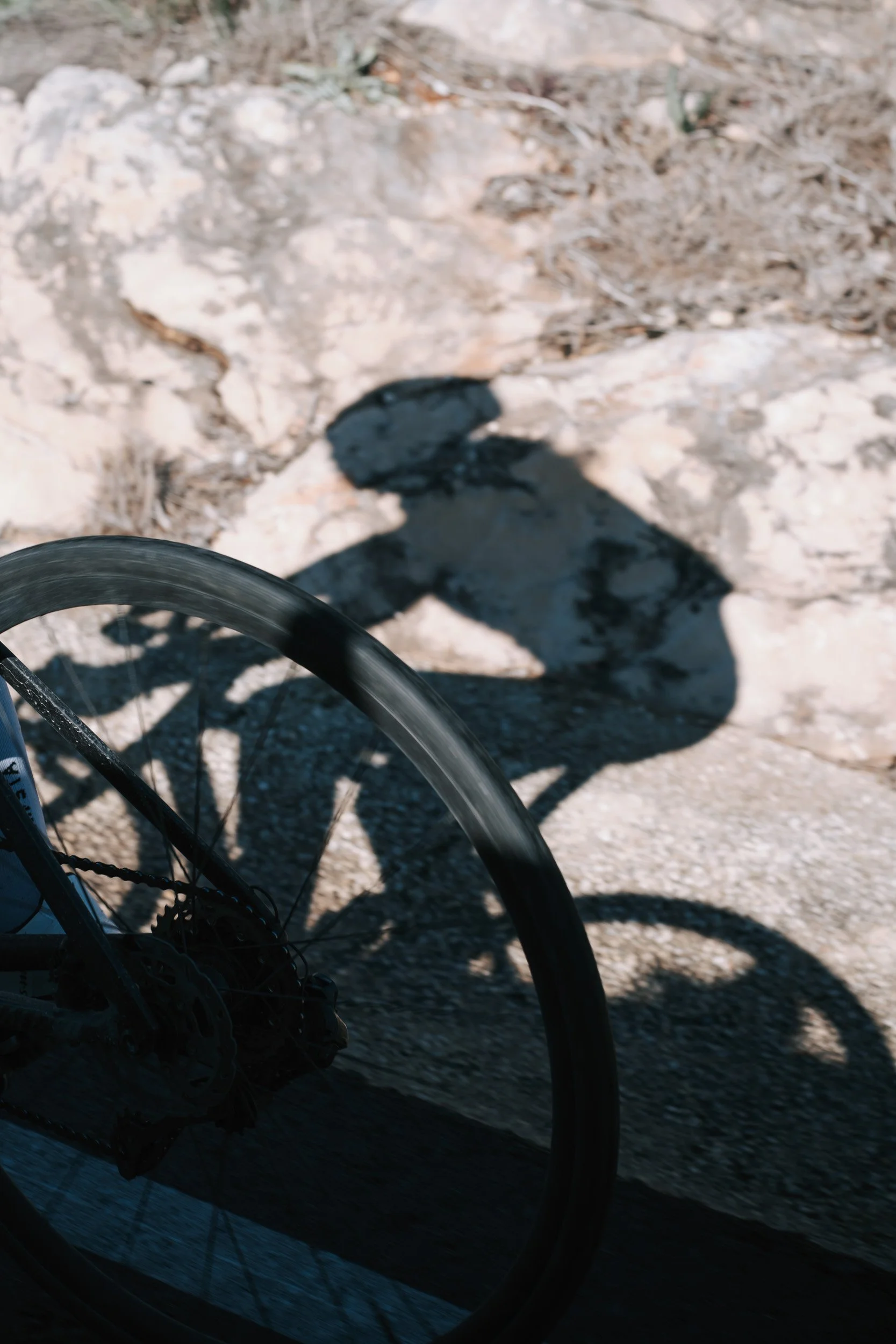 Shadows of a person wearing a baseball cap and sunglasses, standing next to a bicycle, cast on rocky ground.