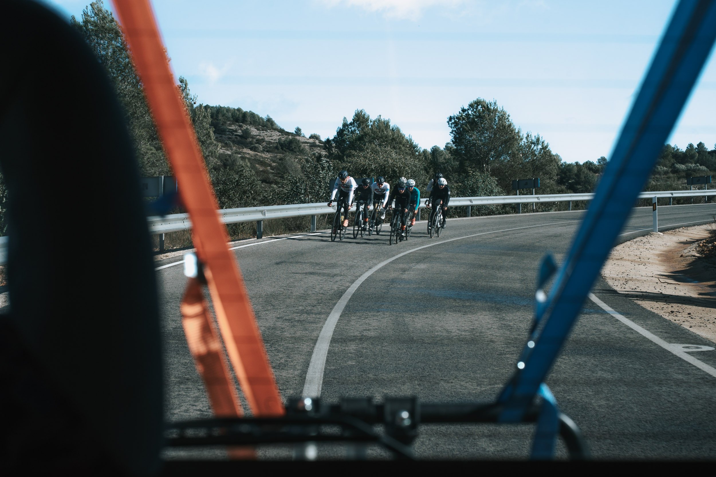 Six cyclists riding on a curved mountain road, seen through the frame of a vehicle.
