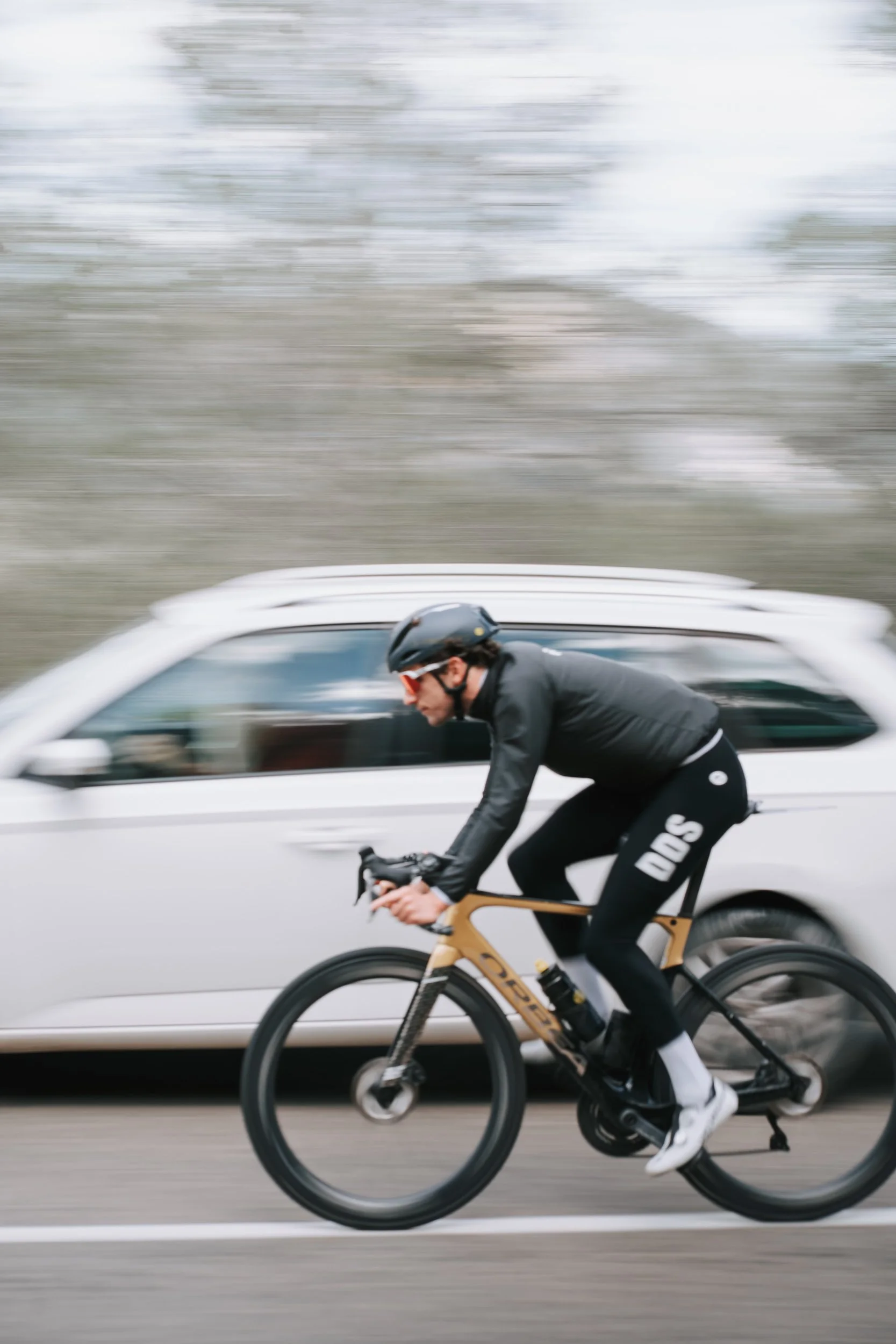 A man wearing a helmet and sunglasses riding a road bike on a street with a moving white vehicle nearby, with blurred background indicating motion.