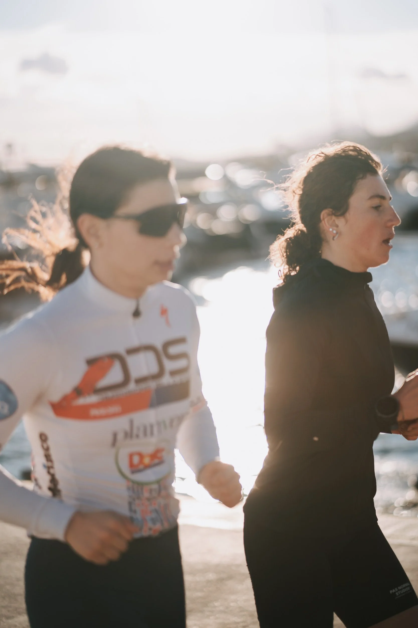 Two women jogging along a waterfront with boats in the background during sunset.