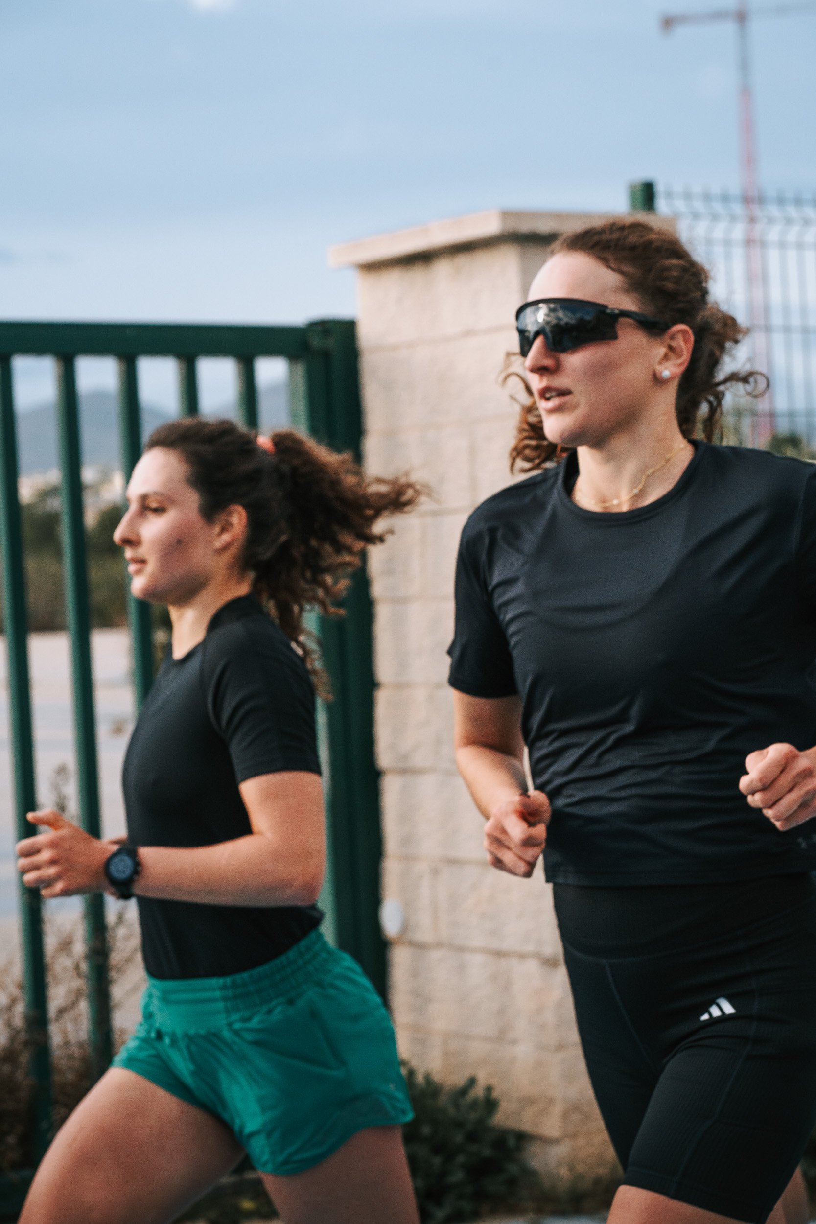 Two women running outdoors near a green iron fence and stone wall, dressed in black athletic clothing, with one wearing sunglasses.
