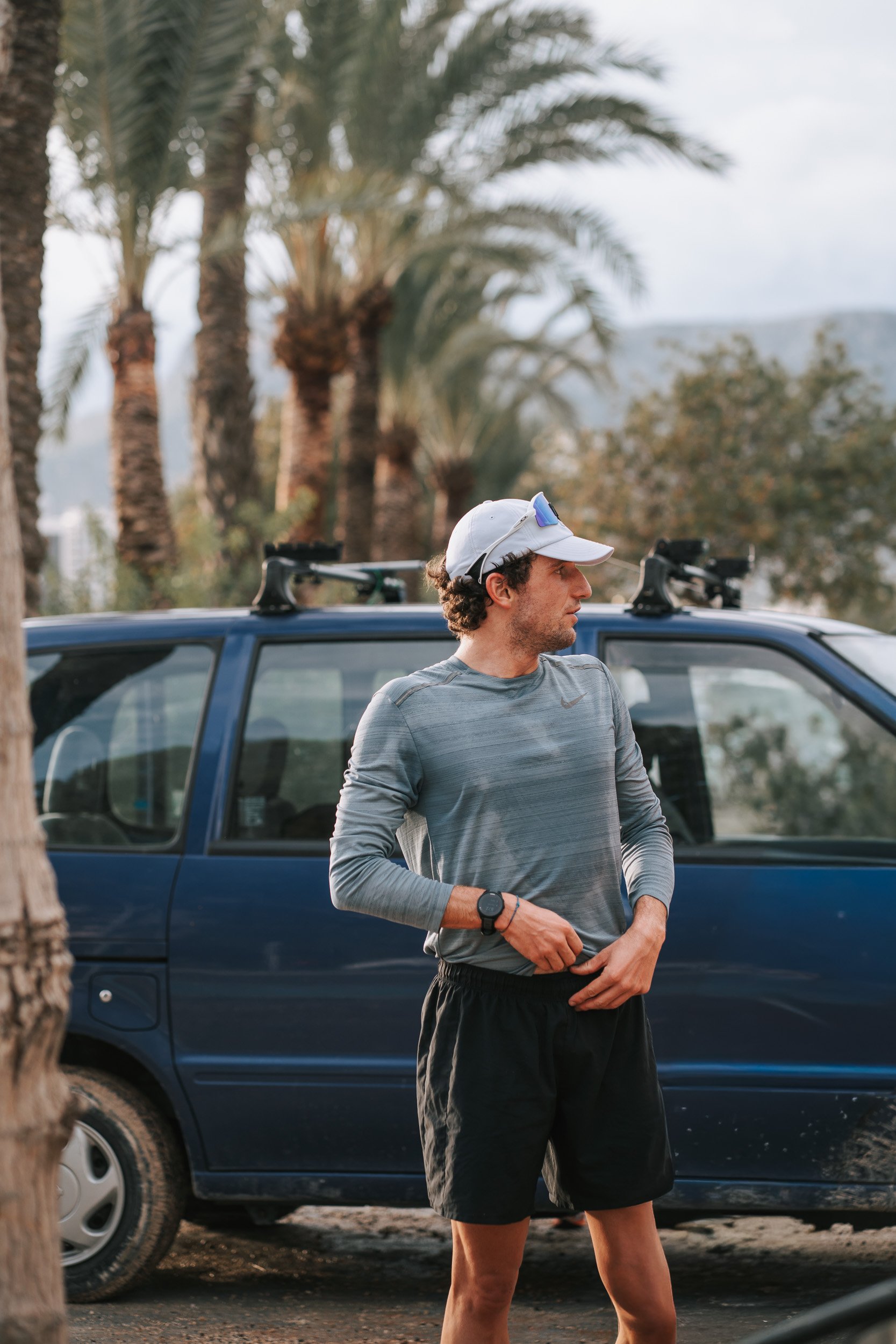 A man standing outdoors next to a blue vehicle, surrounded by palm trees, wearing a gray long-sleeve athletic shirt, black shorts, a white cap, sunglasses, and a wristwatch.