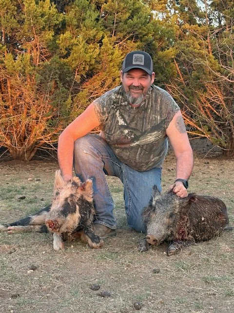 A man kneeling outdoors on dirt terrain with two hunted wild boars, lying on the ground, surrounded by bushes and trees.
