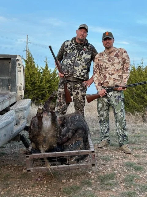 Two men in camouflage clothing standing outdoors behind a wild boar in a metal trap, holding rifles, with a truck and trees in the background.