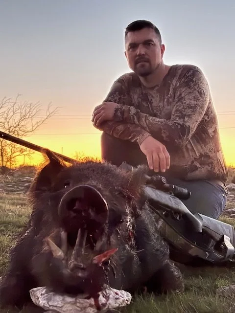 A man in camouflage clothing kneels next to a dead boar with a fishing hook in its mouth during sunset in an outdoor setting.