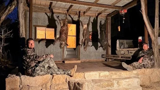 Two men in camouflage clothing sitting on a rustic porch with stone foundation, wooden beams, hunting rifles, and mounted animal hides, at night.