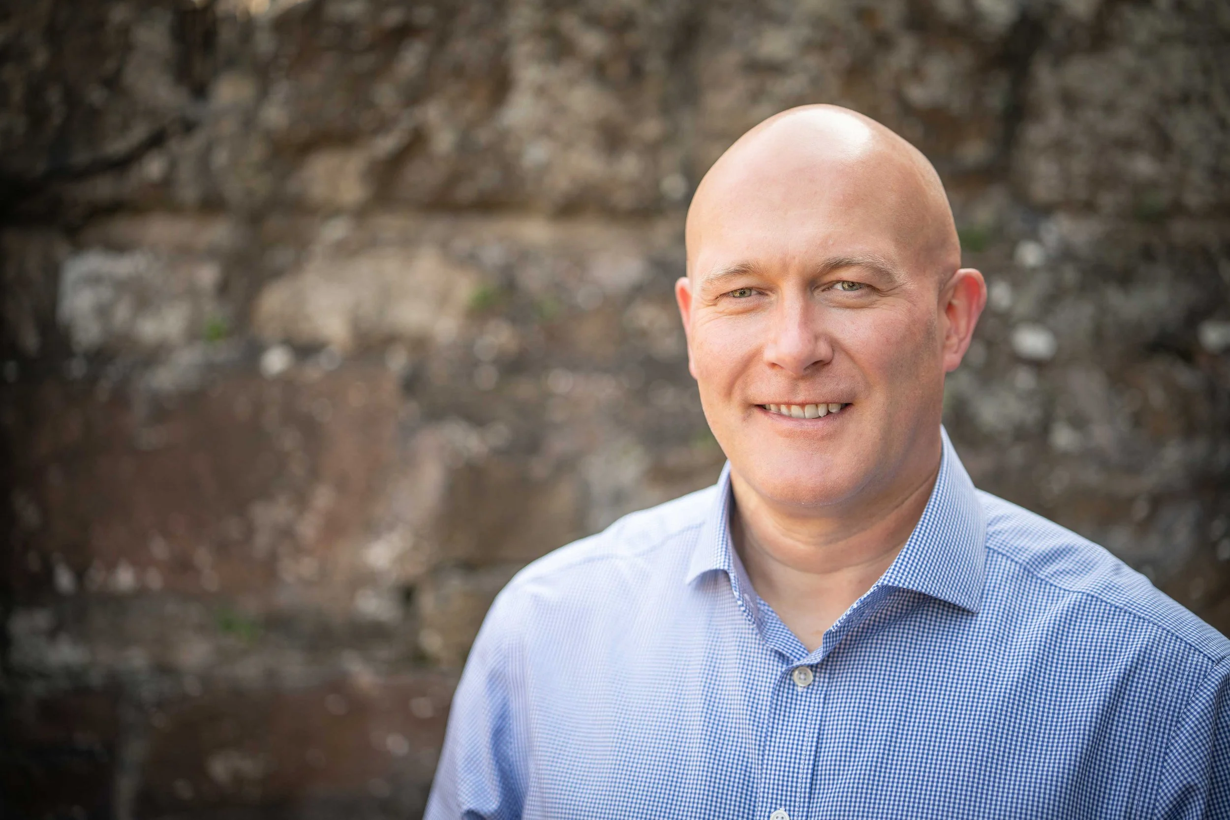 Robert Bell, Bell Financial Planning wearing light blue checkered shirt smiling outdoors with a blurred stone wall background.