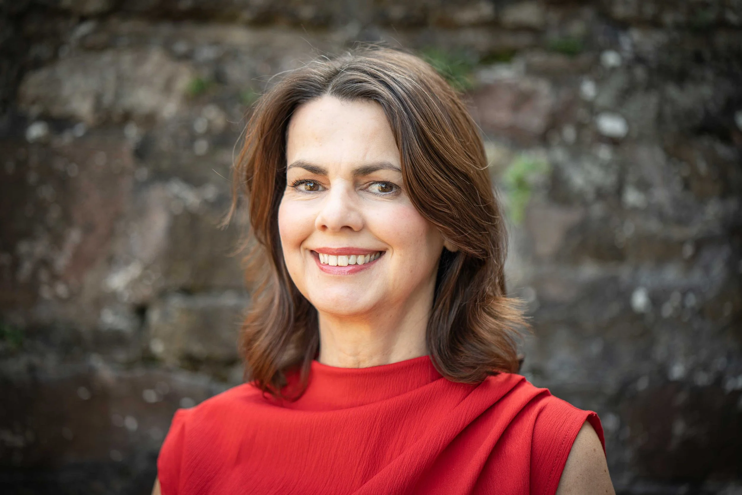 Sandra Bell wearing a red sleeveless top, smiling in front of a stone wall.