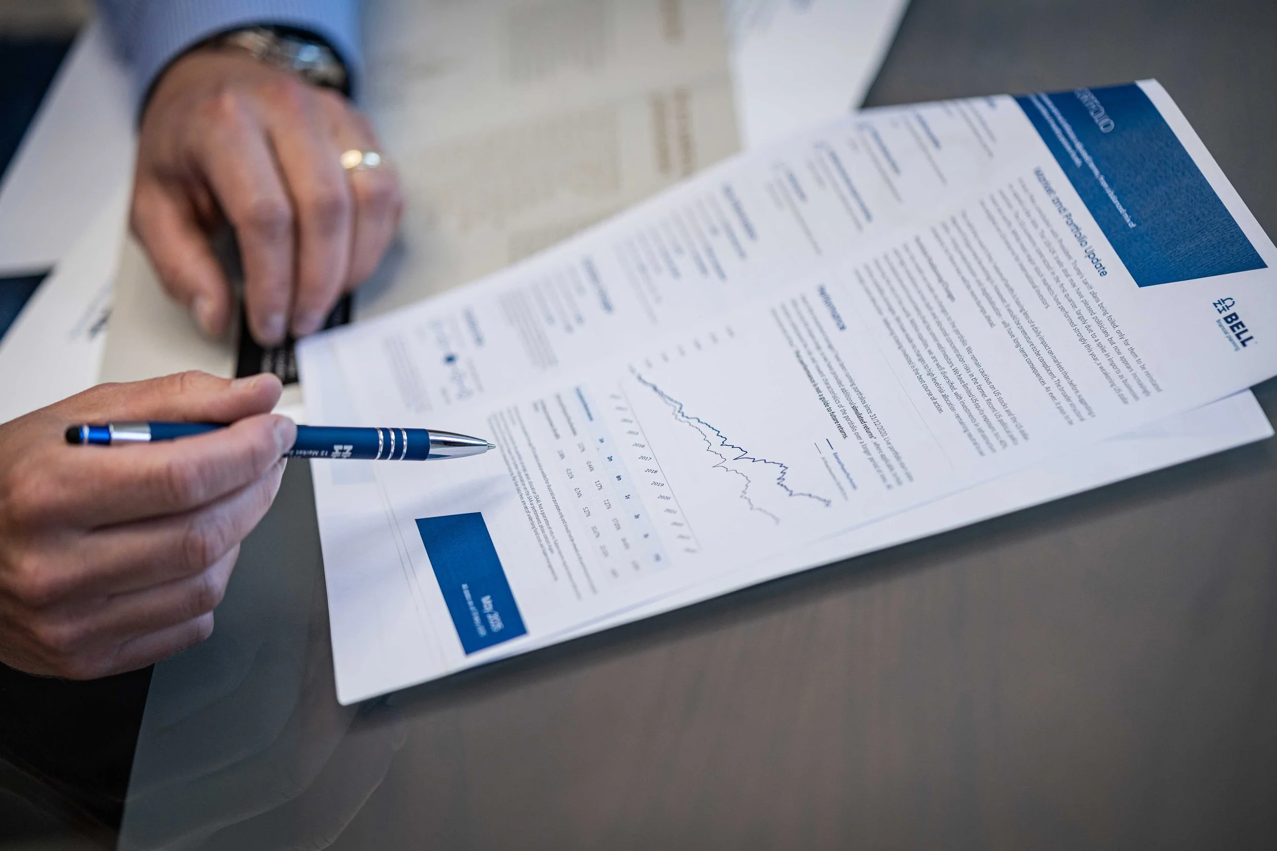Close-up of a person pointing at a financial report with a pen, showing graphs and charts on the document.