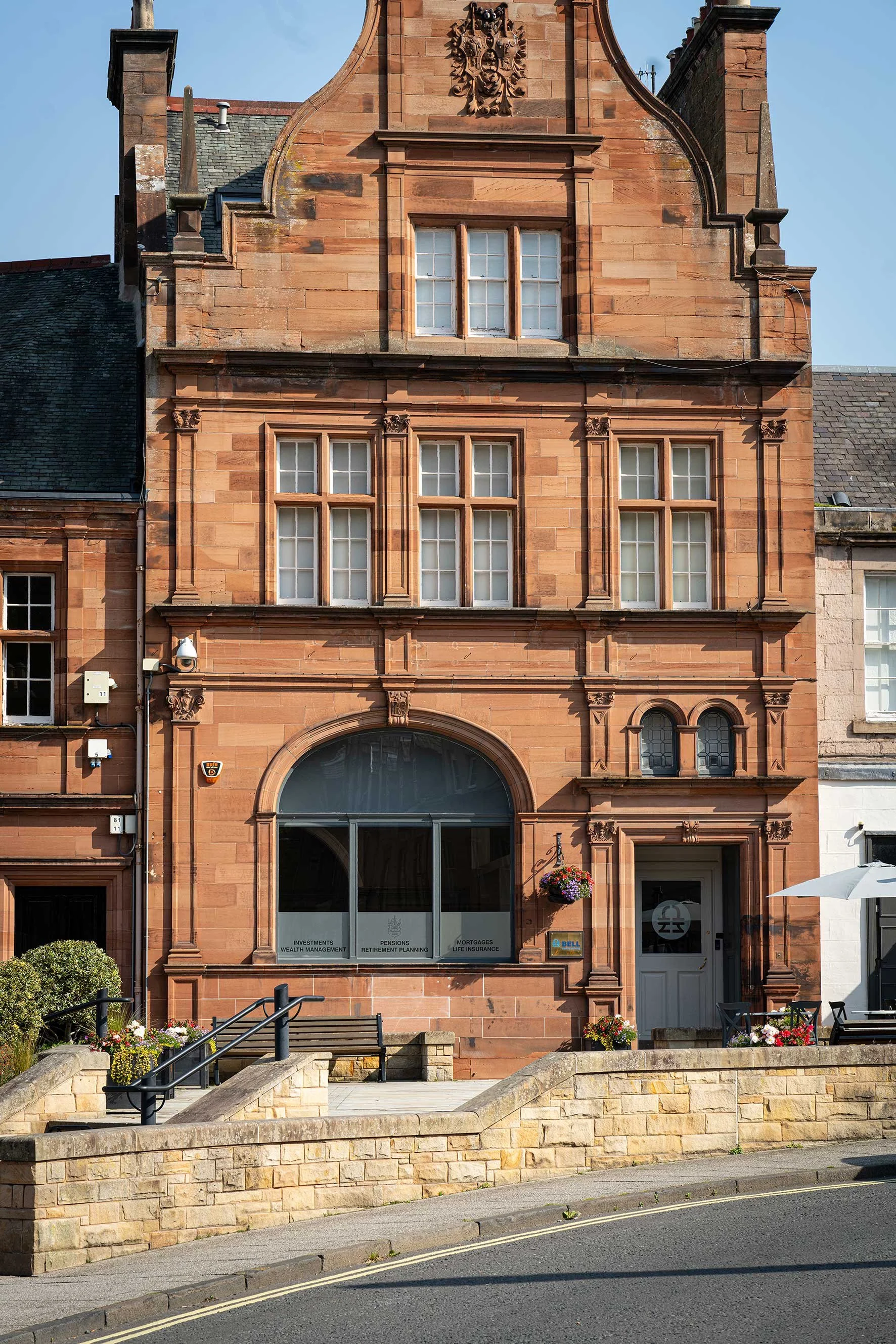 Bell Financial Planning. A historic red brick building with ornate architectural details, multiple windows, a large arched window displaying investment and retirement planning signs, and a white door surrounded by flowers and outdoor furniture.