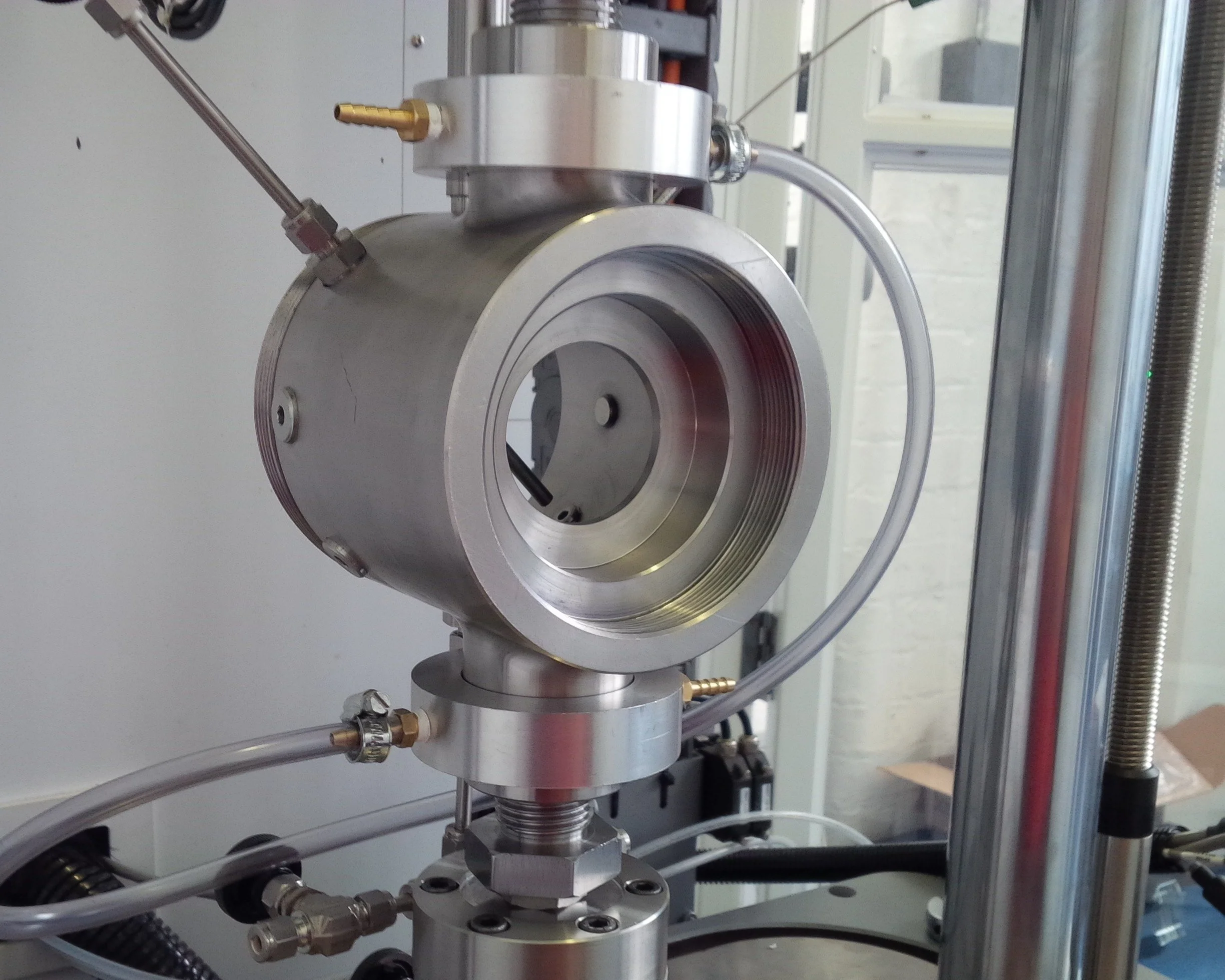 Close-up of a scientific or industrial apparatus made of metal with various parts, including fittings, tubing, and a large circular component, in a laboratory or manufacturing setting.