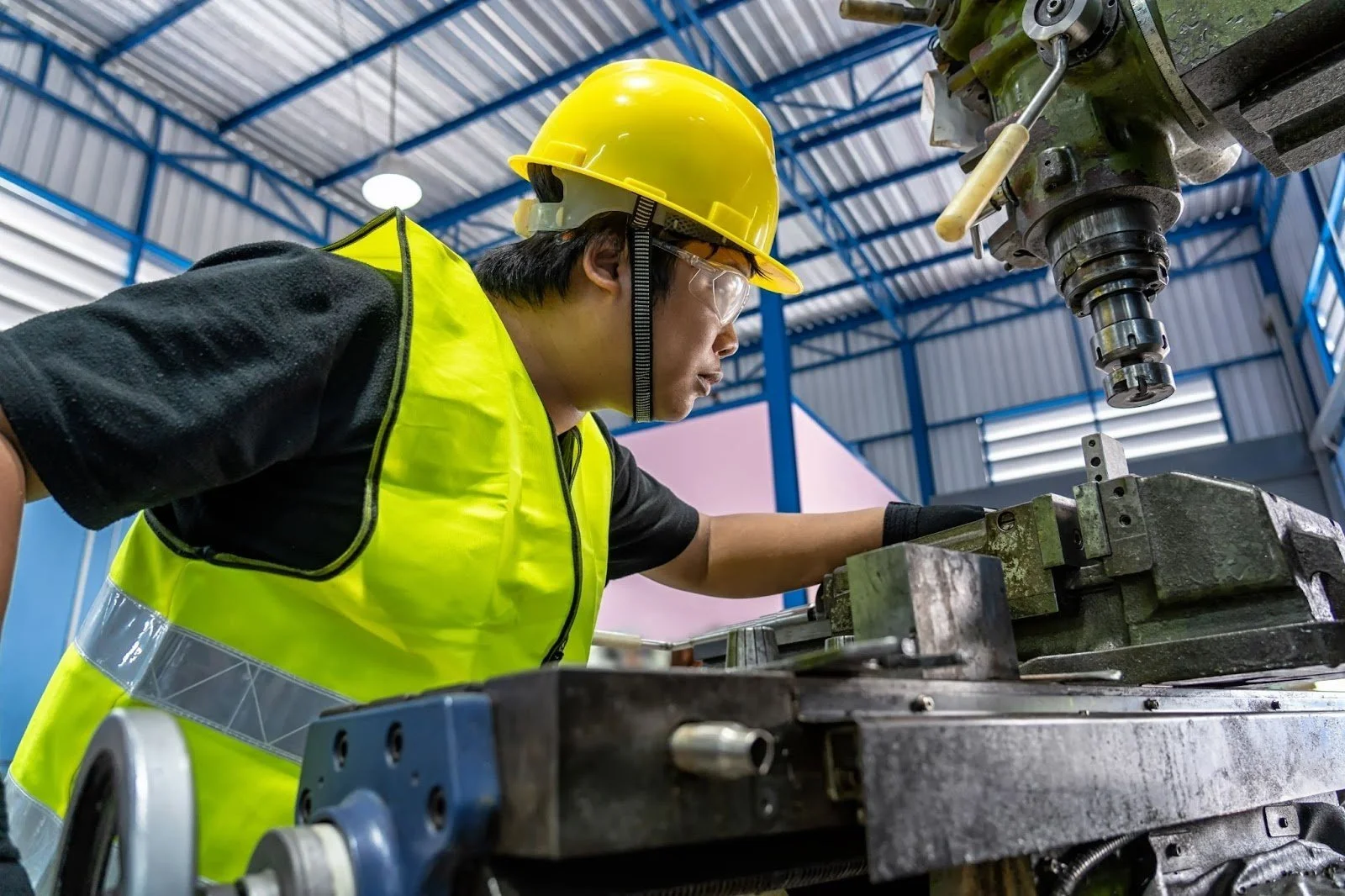 A worker in a yellow safety helmet and high-visibility vest operating a large industrial machine in a factory or workshop.