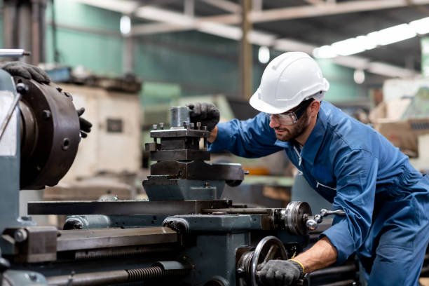 A man wearing a white safety helmet and safety glasses operating a large industrial lathe machine in a factory setting.