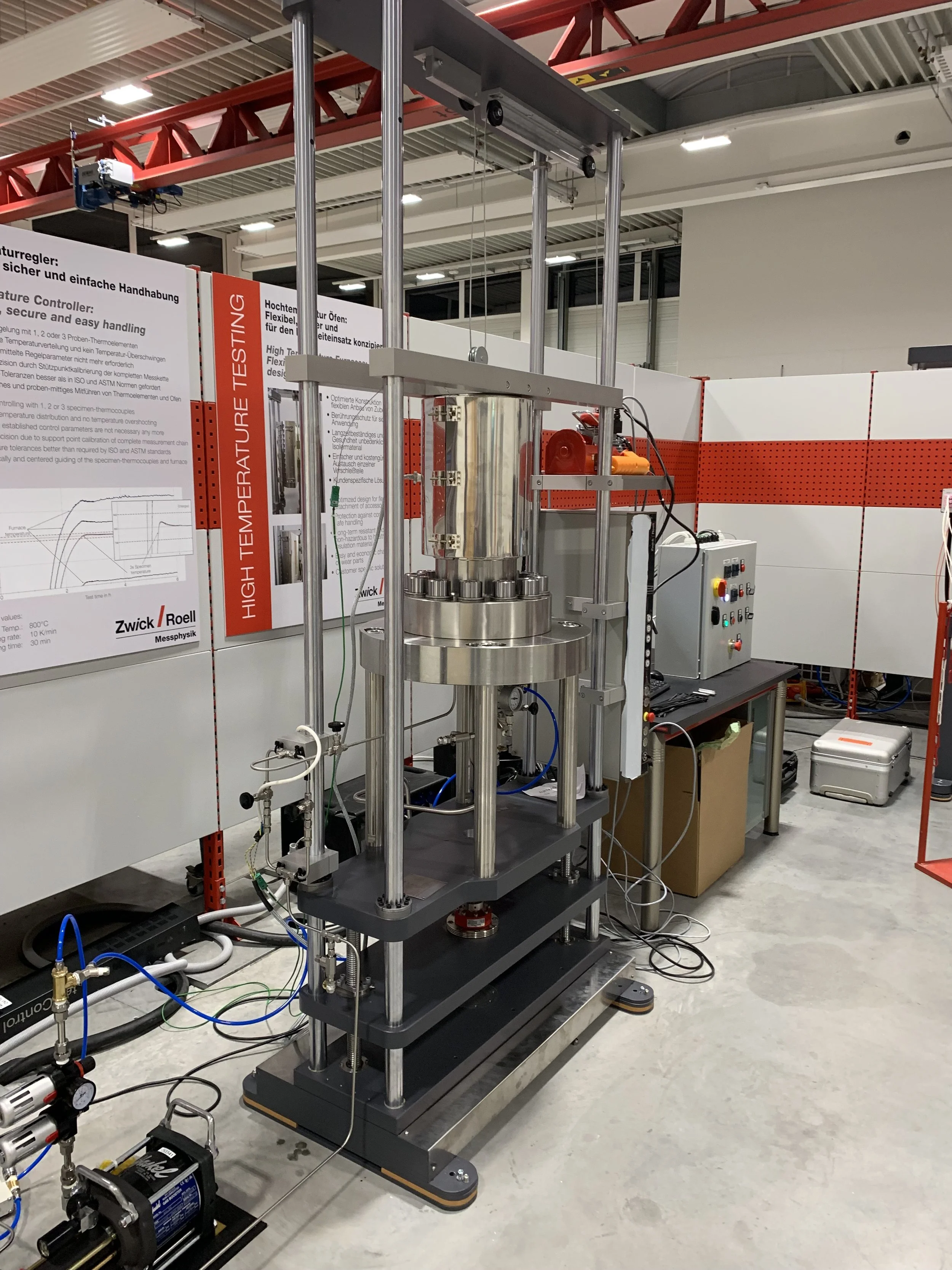 Laboratory setup with scientific equipment, including a vertical cylindrical metallic apparatus with a red motor on the side, connected to various tubes, wires, and control panels, and informational posters in the background.