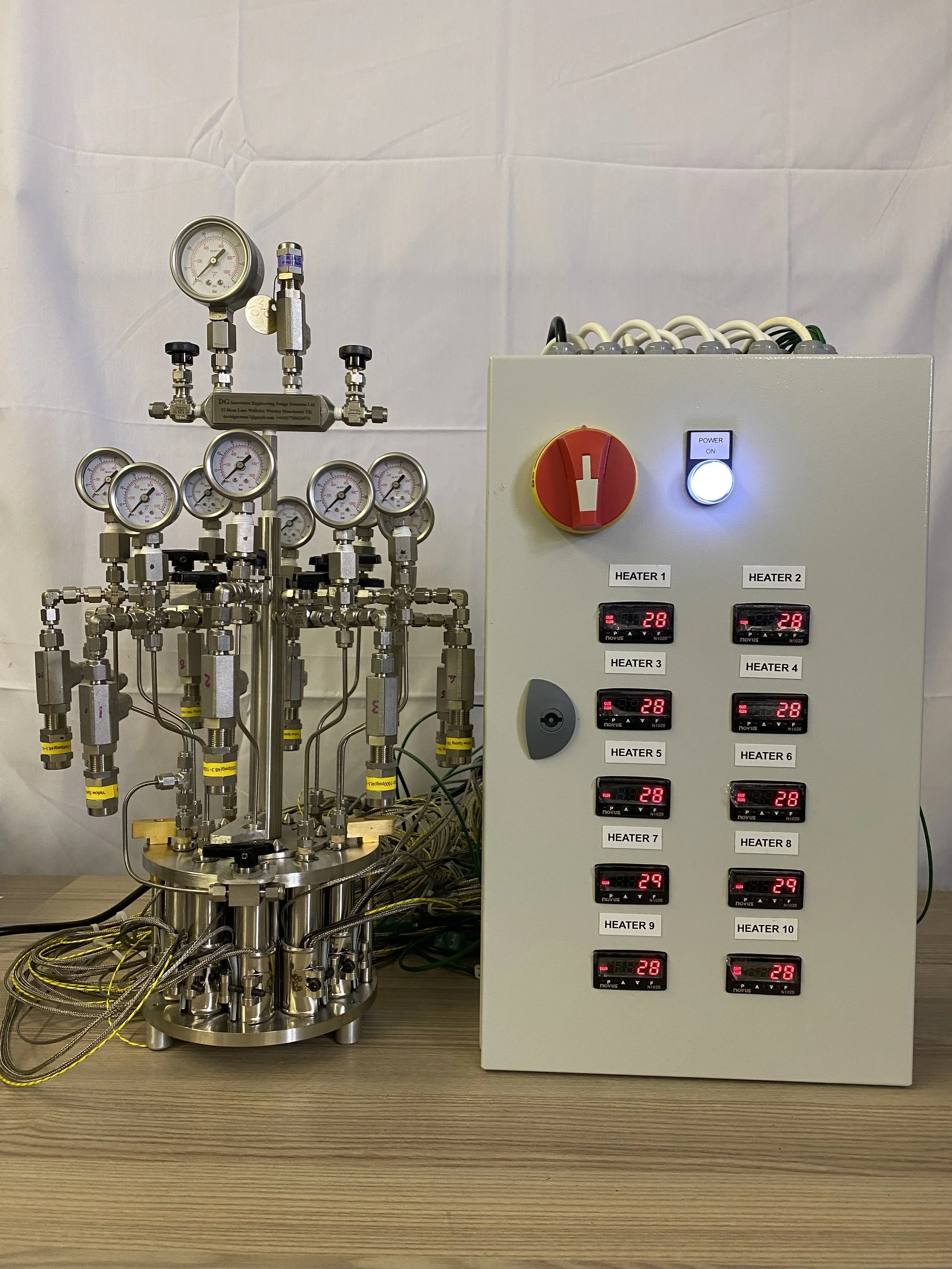Laboratory equipment with various gauges and wires on a wooden table, featuring a control panel with digital temperature displays and switches labeled for different heaters.