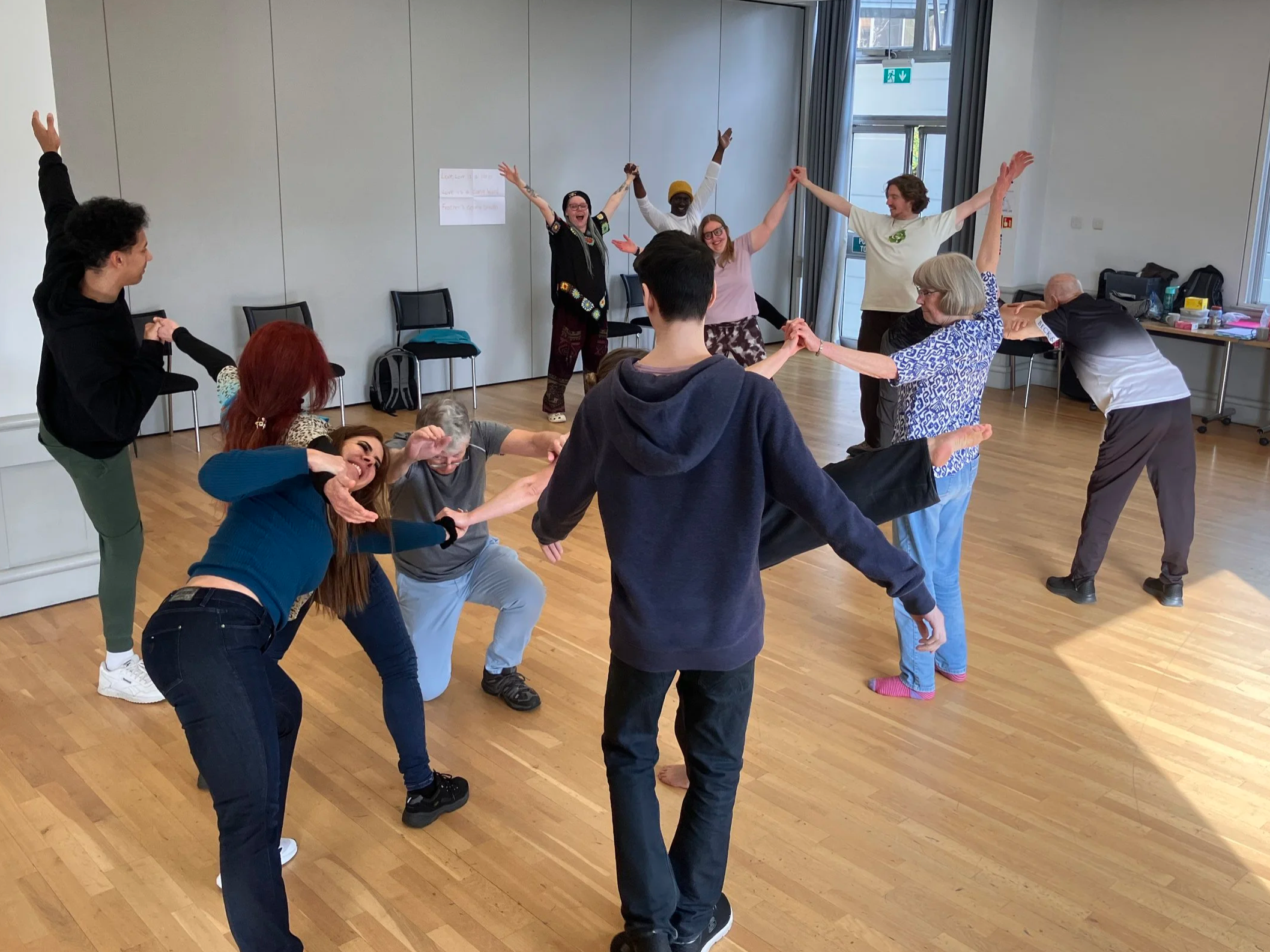 a group of people spread out across a bright room. Some are standing, some are kneeling. Some people have arms and legs outstretched to connect with others. People are smiling and having fun.