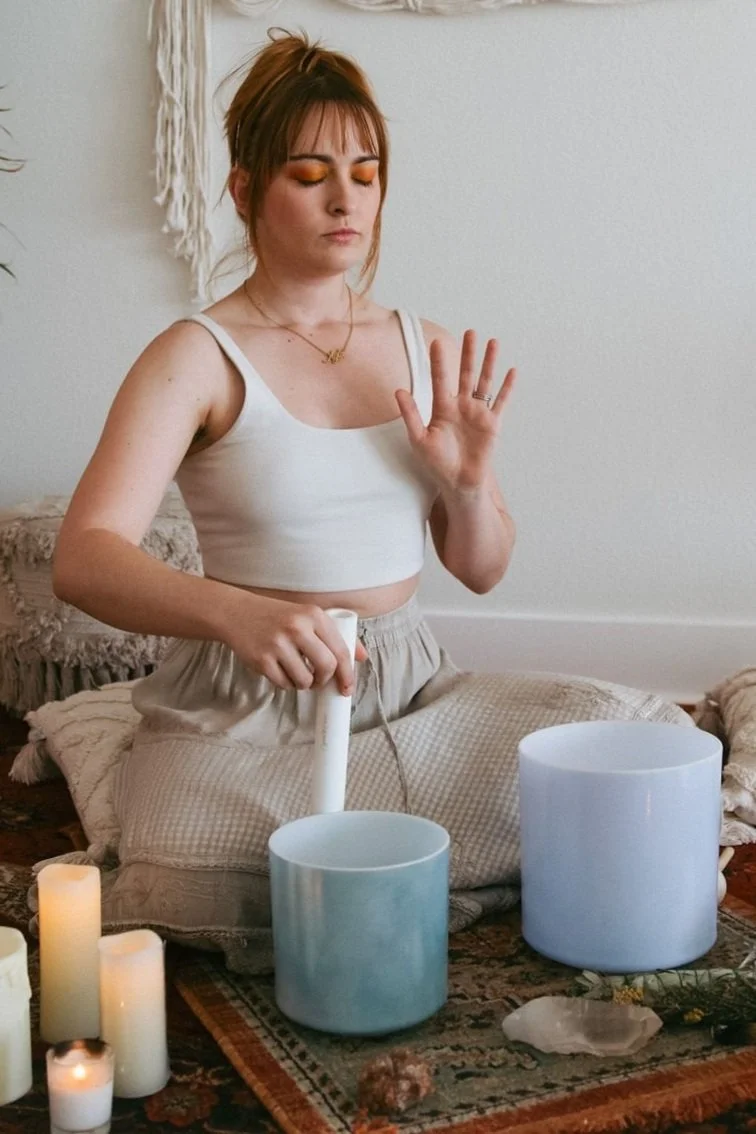 A young woman with short light brown hair and orange eyeshadow is sitting cross-legged on a rug with candles and large singing bowls in front of her. She is holding a mallet in one hand and raising her other hand with her eyes closed, appearing to meditate or perform a sound healing session.