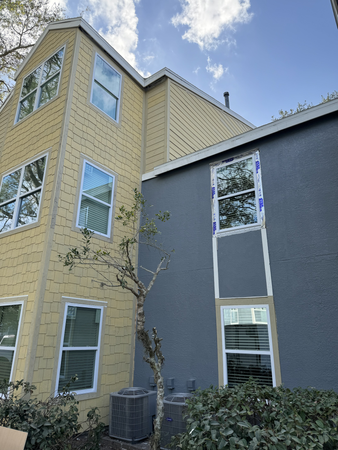 Exterior view of a two-story building with yellow siding and grey stucco sections, featuring multiple windows and surrounded by shrubs. An air conditioning unit is visible beside the building, with a partially cloudy sky overhead.