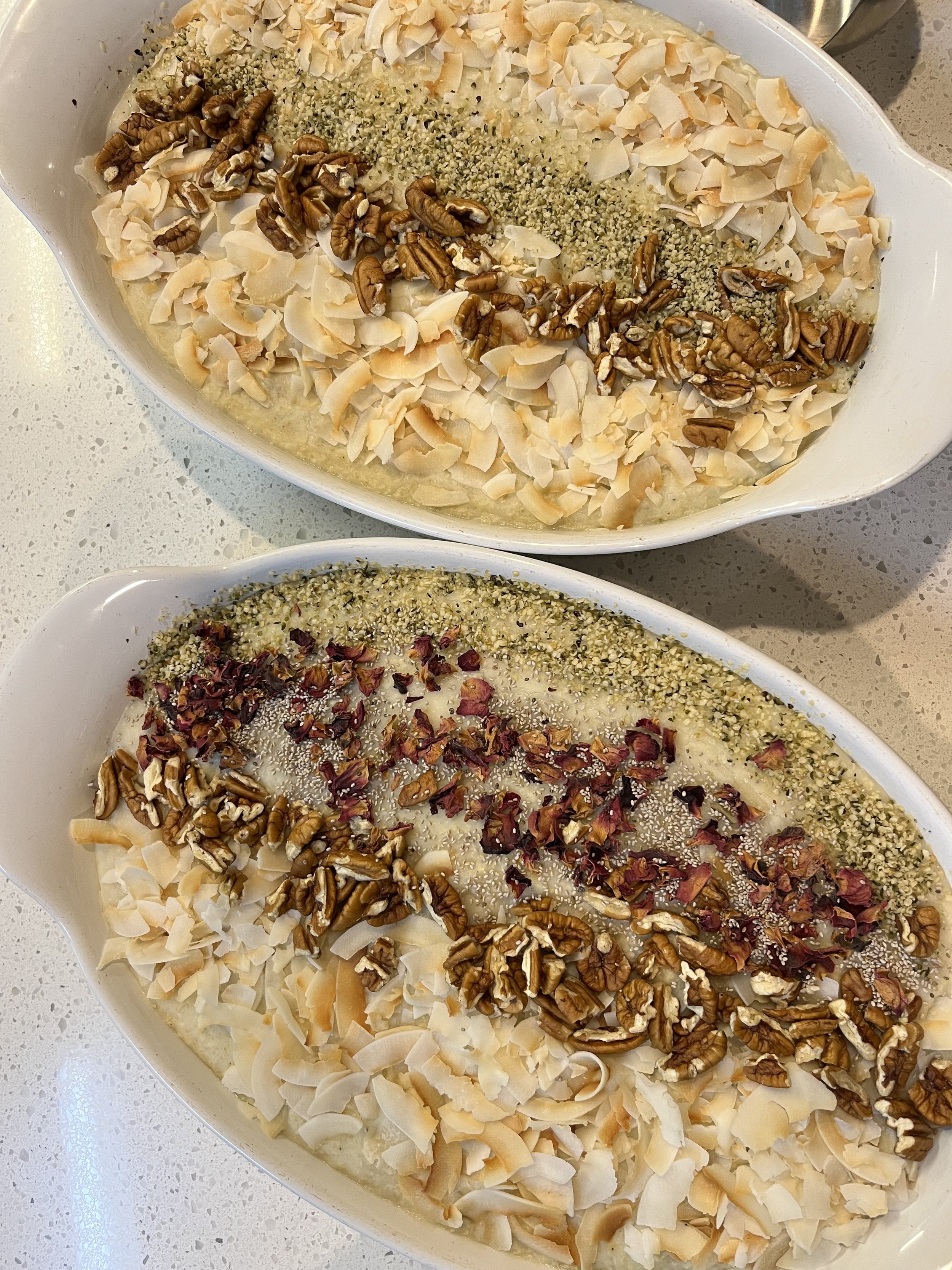 Two bowls of oatmeal topped with shredded coconut, pecans, hemp seeds, and rose petals on a light-colored countertop.