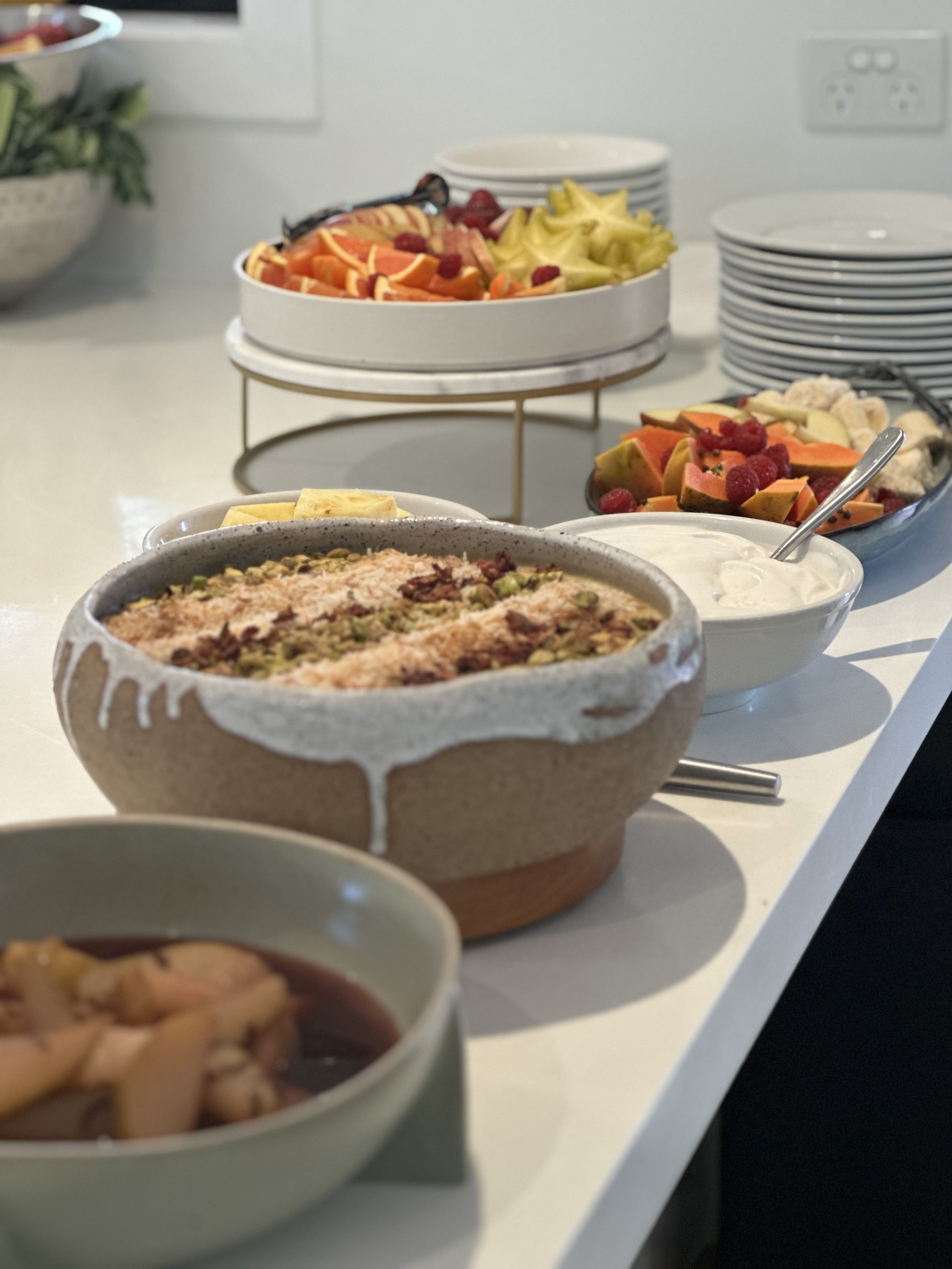 A table with dishes of fruit salad, sliced fruits including berries and starfruit, a ceramic bowl with baked oatmeal or casserole, and a bowl of fruit compote. Plates are stacked in the background.